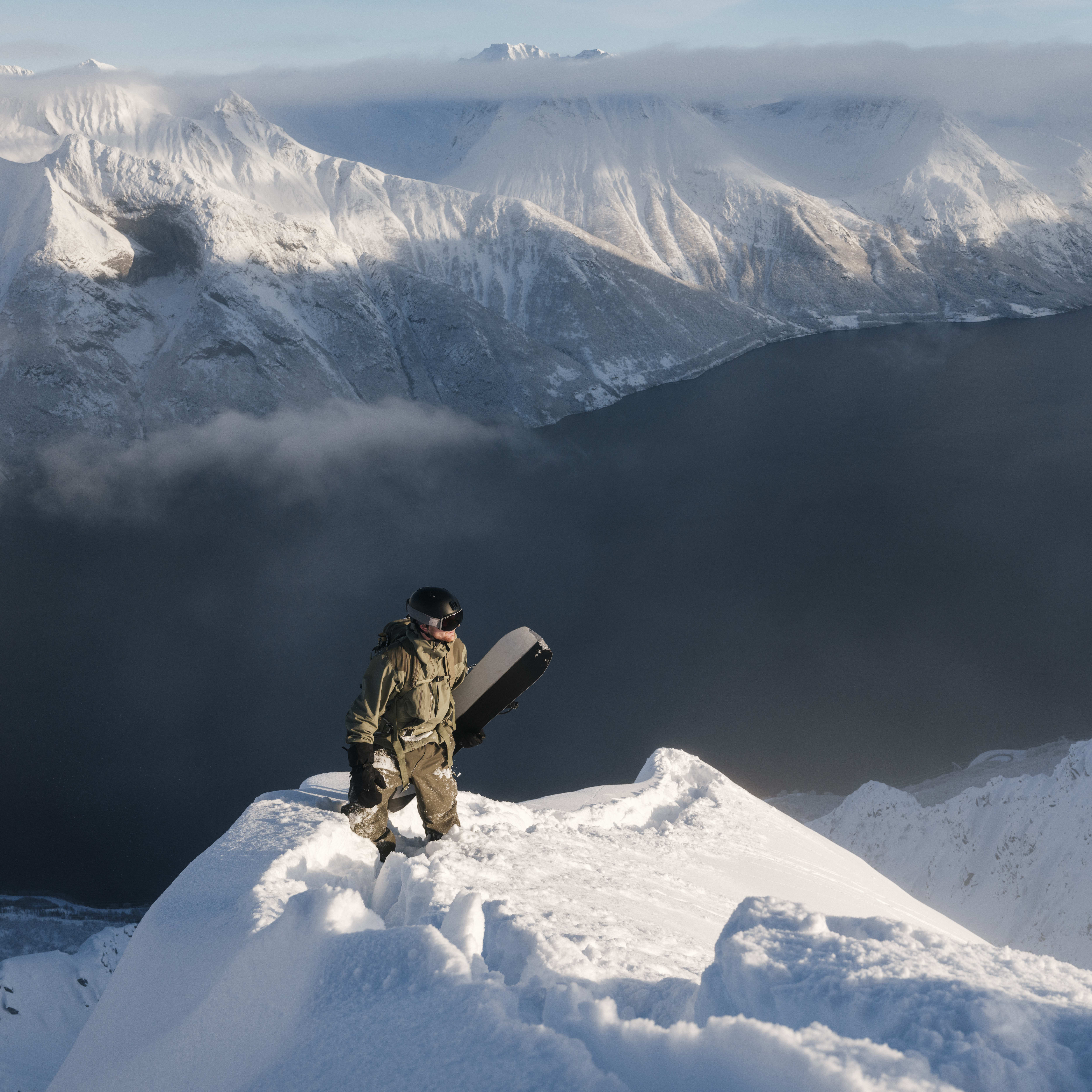 A snowboarder in Norrøna gear stands atop a snowy mountain peak with a fjord and mountain range in the background.