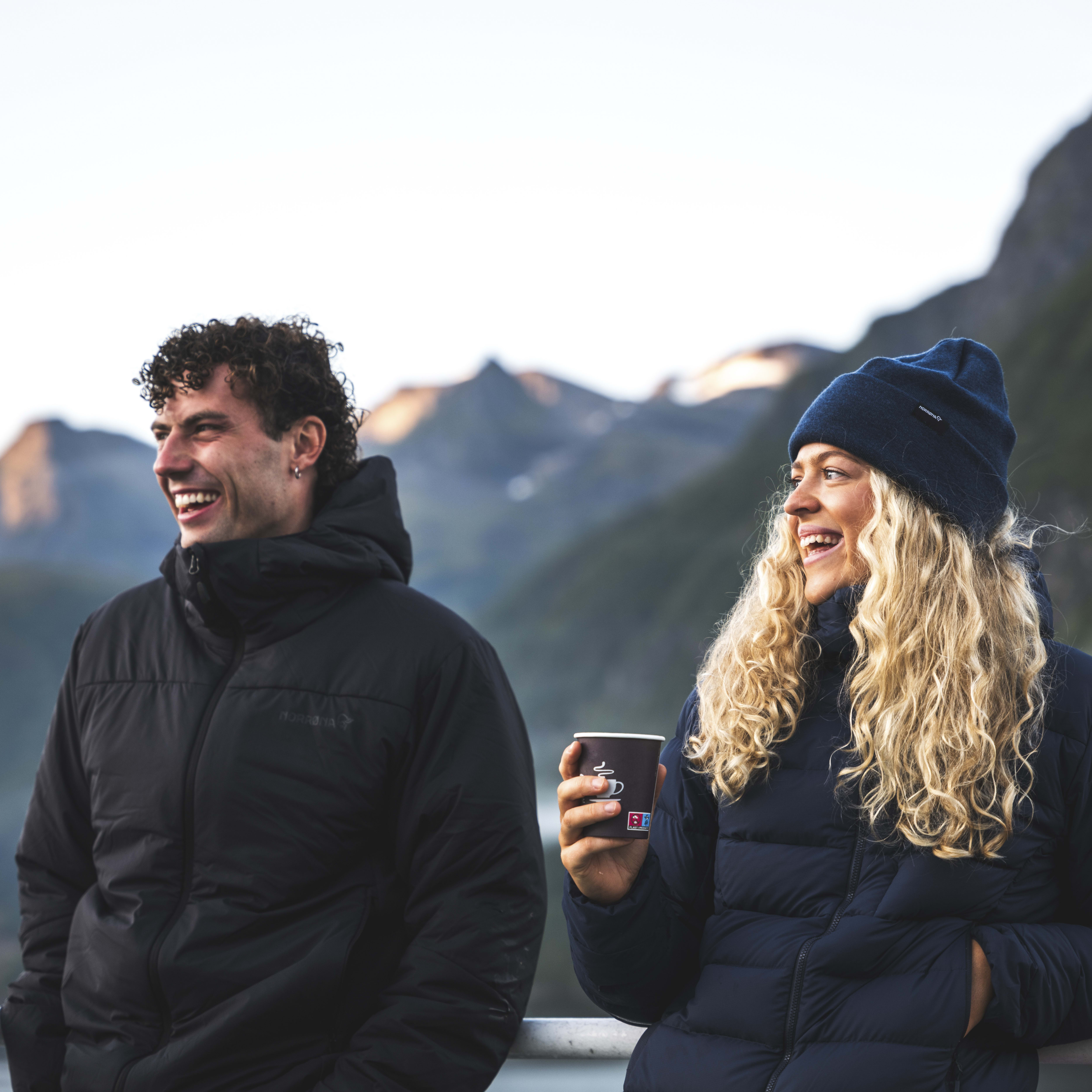 Two smiling people stand outside. The woman wears a blue Norrøna beanie and jacket, and holds a paper coffee cup. The man wears a black Norrøna jacket. Mountains are in the background.