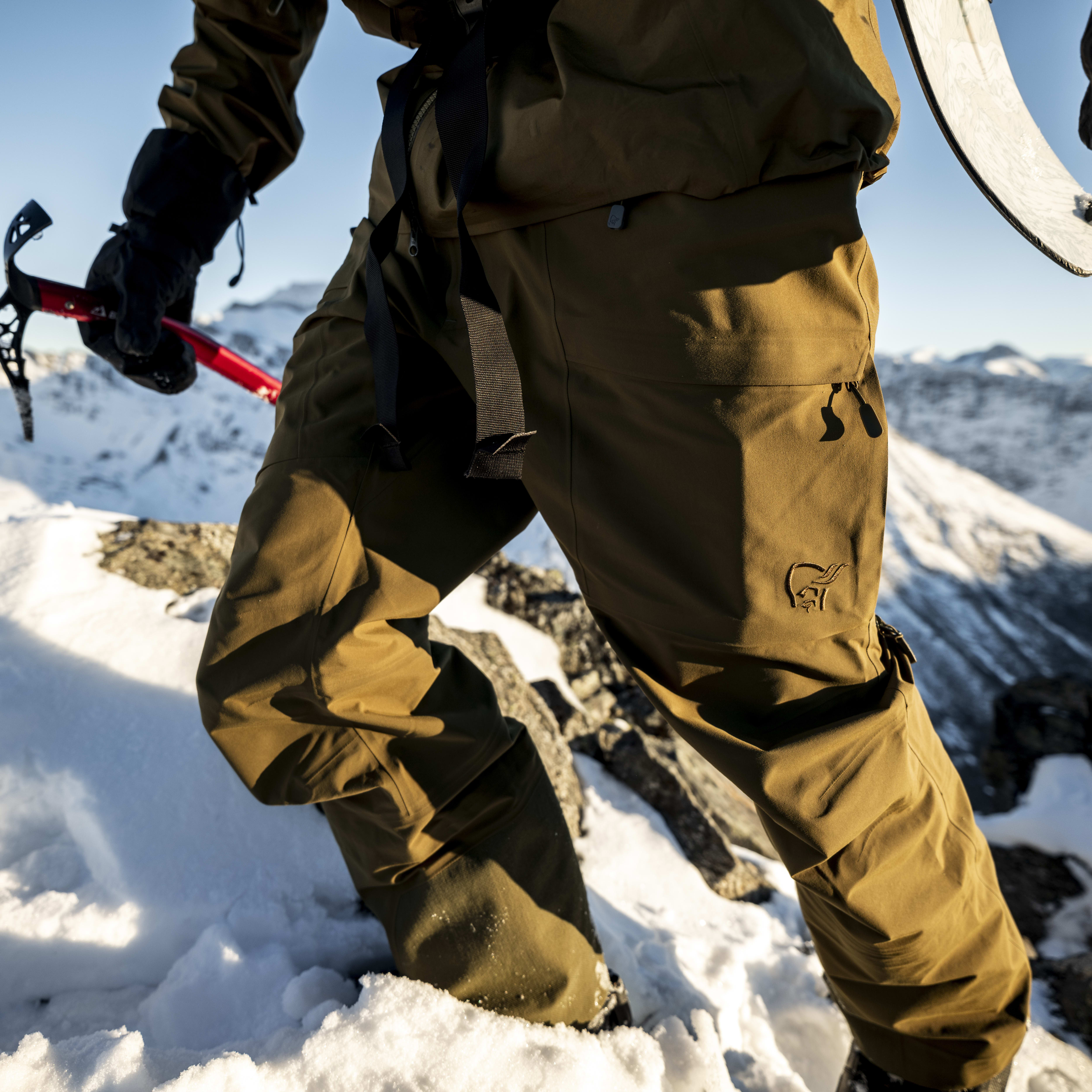 Close-up of a climber with an ice axe, ascending a snowy mountain in Norrøna pants. Skis visible, snowy peaks in the background.