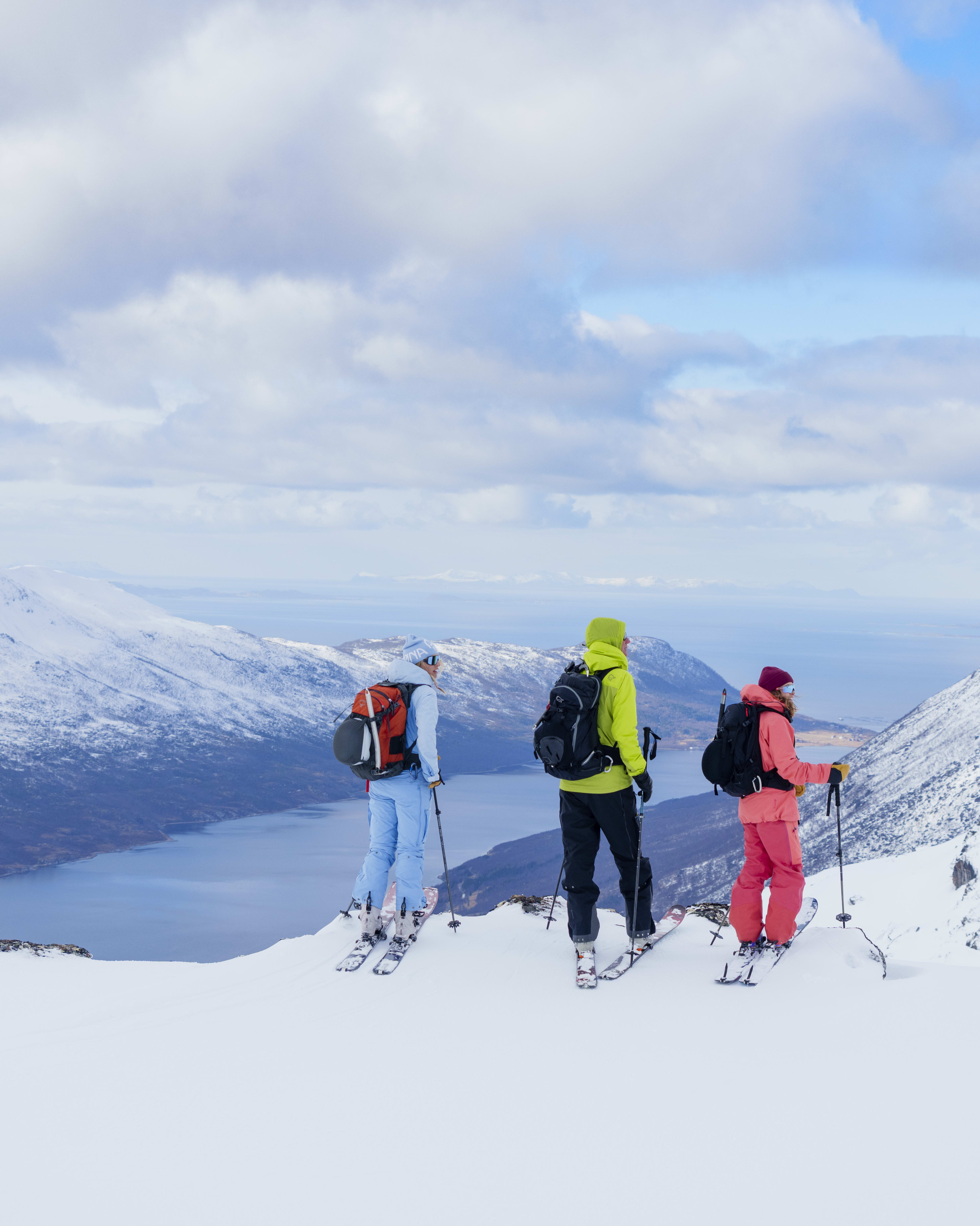 Three skiers in Norrøna gear stand atop a snowy mountain, overlooking a fjord and snowy peaks under a partly cloudy sky.