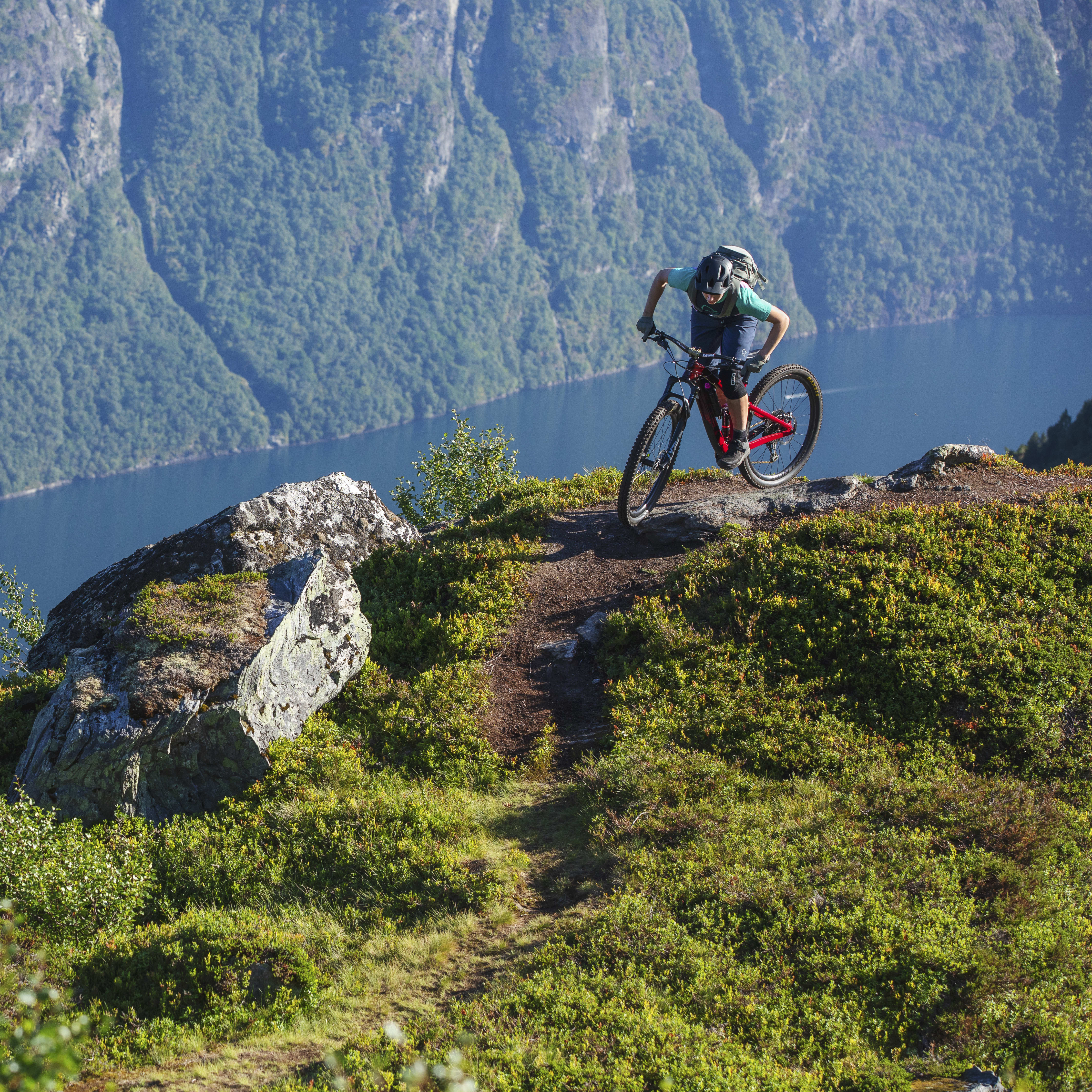 A mountain biker in Norrøna clothing rides a red bike on a trail overlooking a blue fjord with a boat and steep mountains in the background.