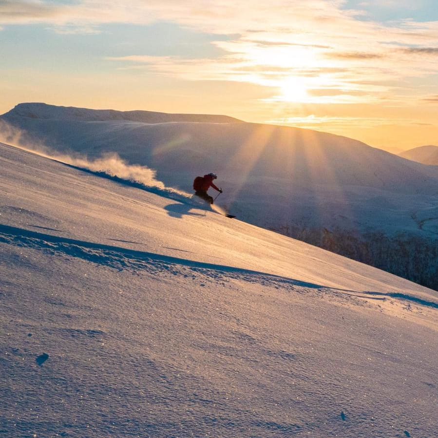 A skier wearing Norrøna clothing, descends a snowy mountain at sunset. Blue sky with wispy clouds.