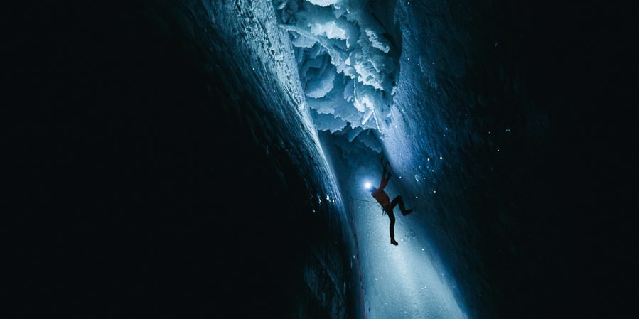 A climber ascends a dark ice cave, illuminated by a headlamp. The climber is wearing Norrøna gear. 
