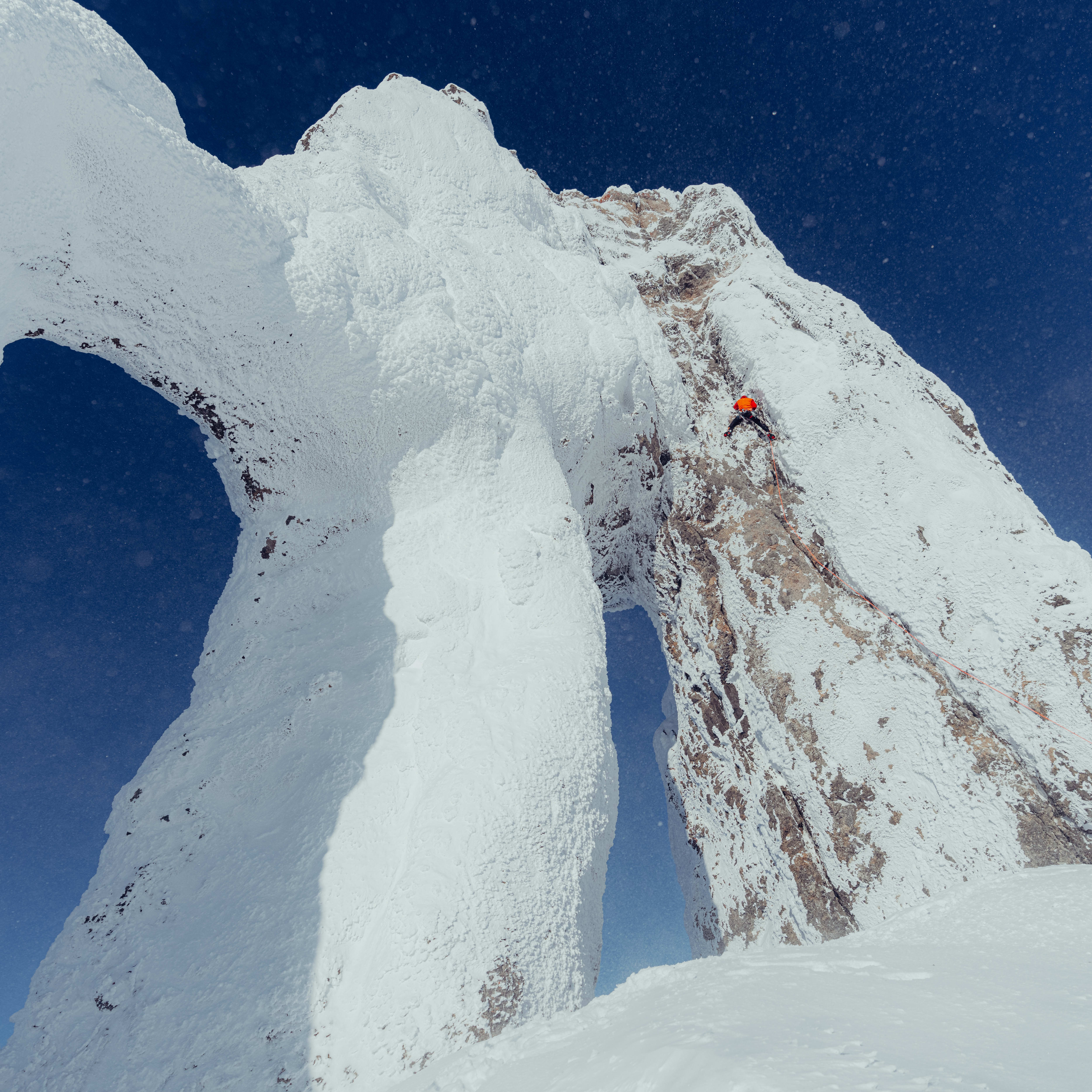 A climber in an orange jacket scales a snow-covered mountain peak against a blue sky. 
