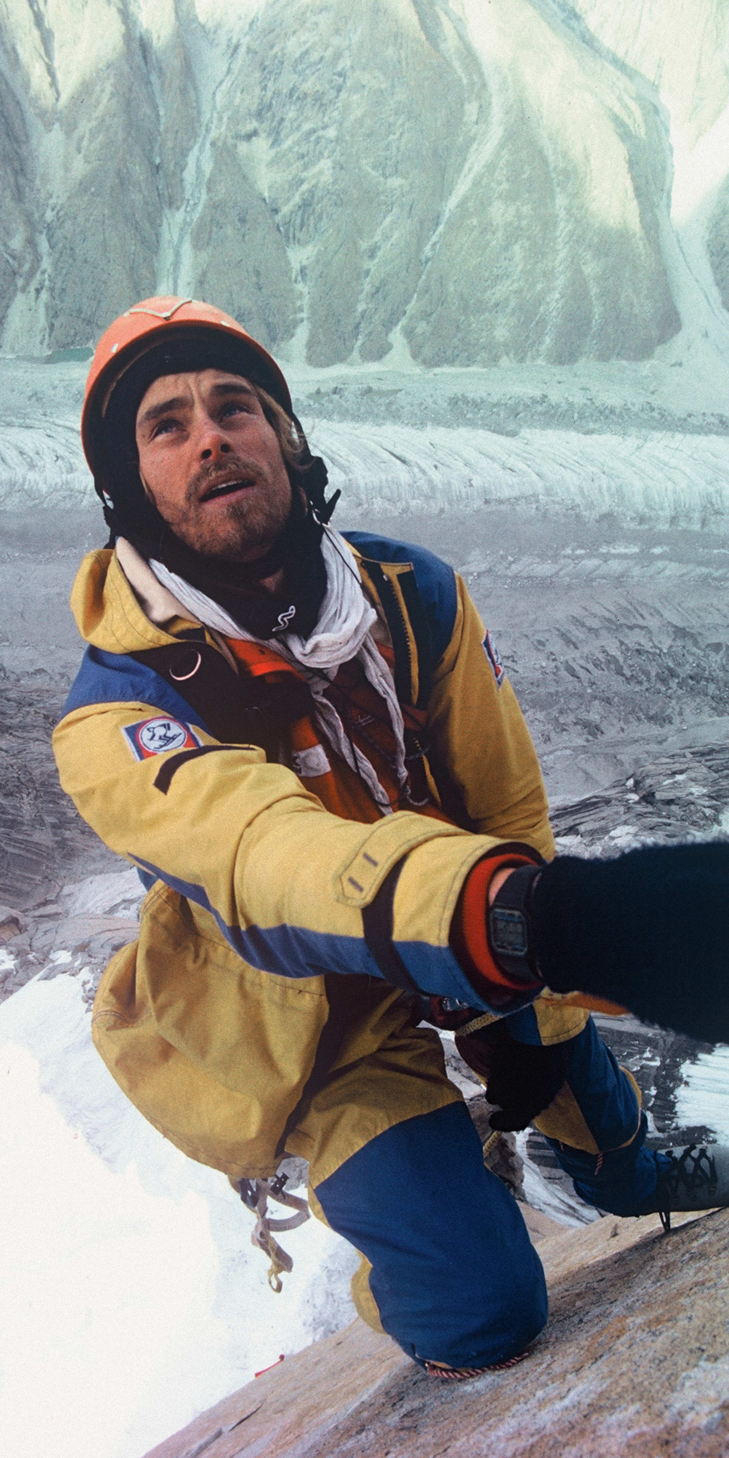 A man wearing a yellow and blue jacket, helmet, and gloves is rock climbing with a yellow rope. He is looking up at the snow covered mountain face.  He appears to be using a belay device and climbing toward a snow covered ridge.