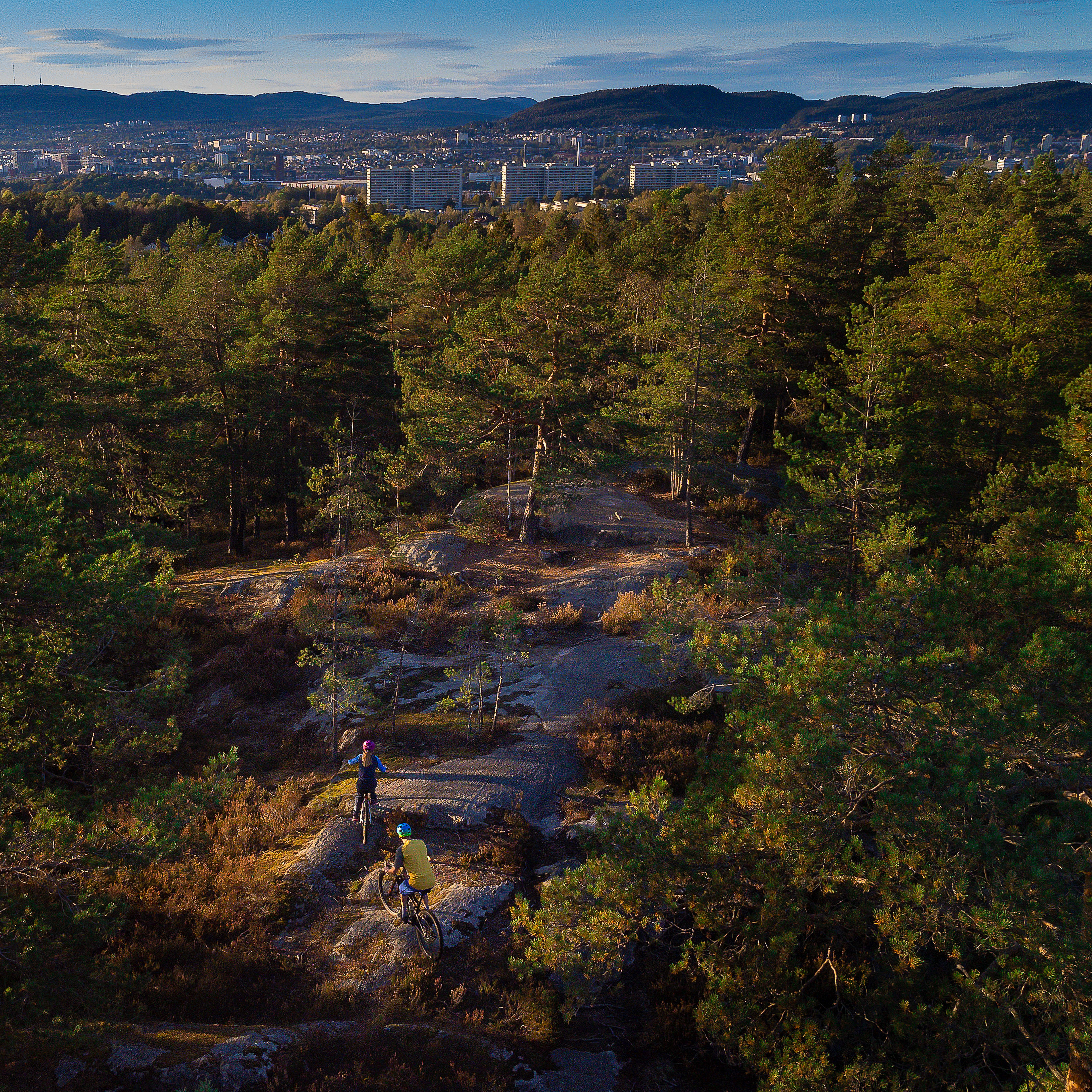 Aerial view of three people mountain biking on a trail in a forest. The city is visible in the background.
