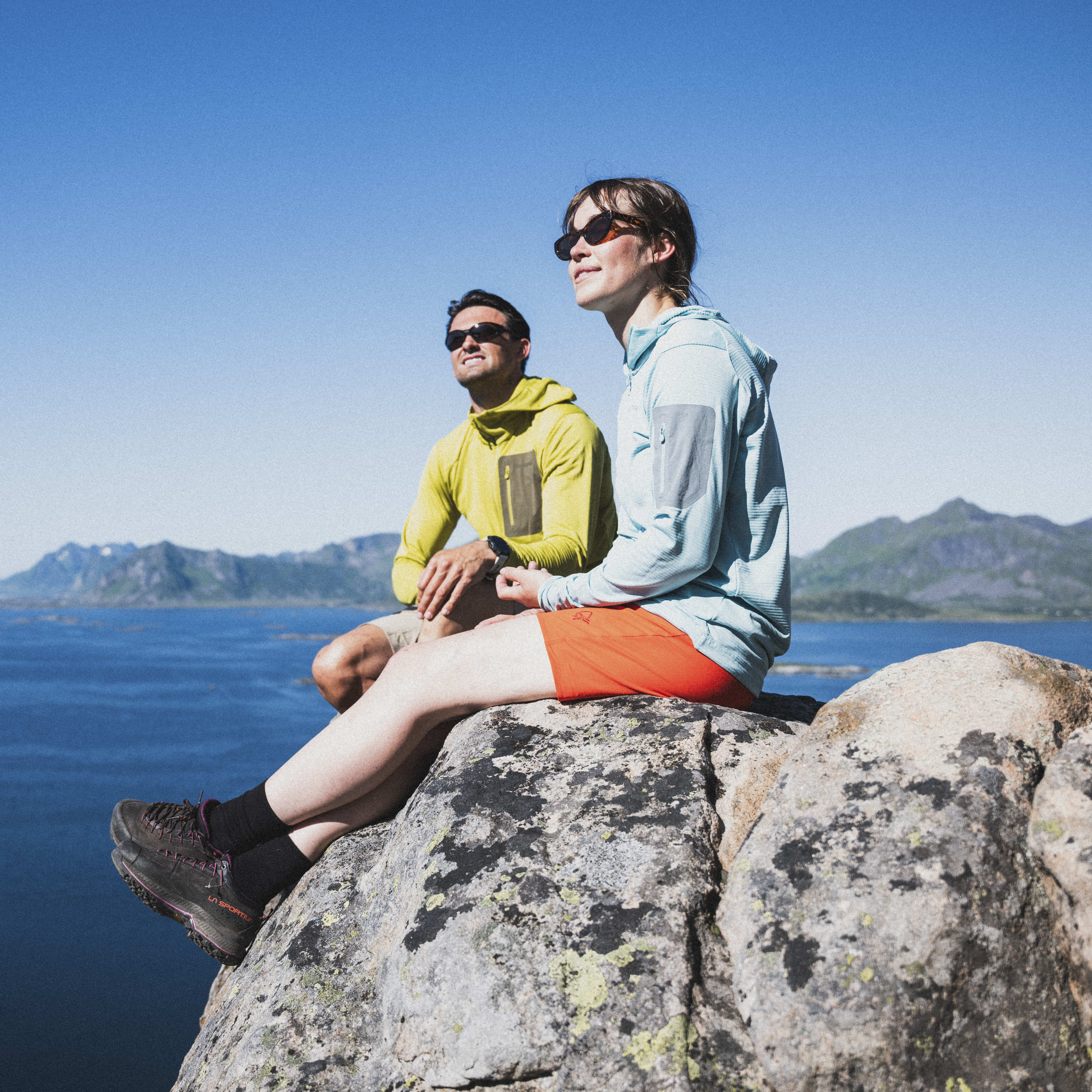 Two people in Norrøna outdoor clothing sit on a rock overlooking a blue ocean and mountains under a bright blue sky.
