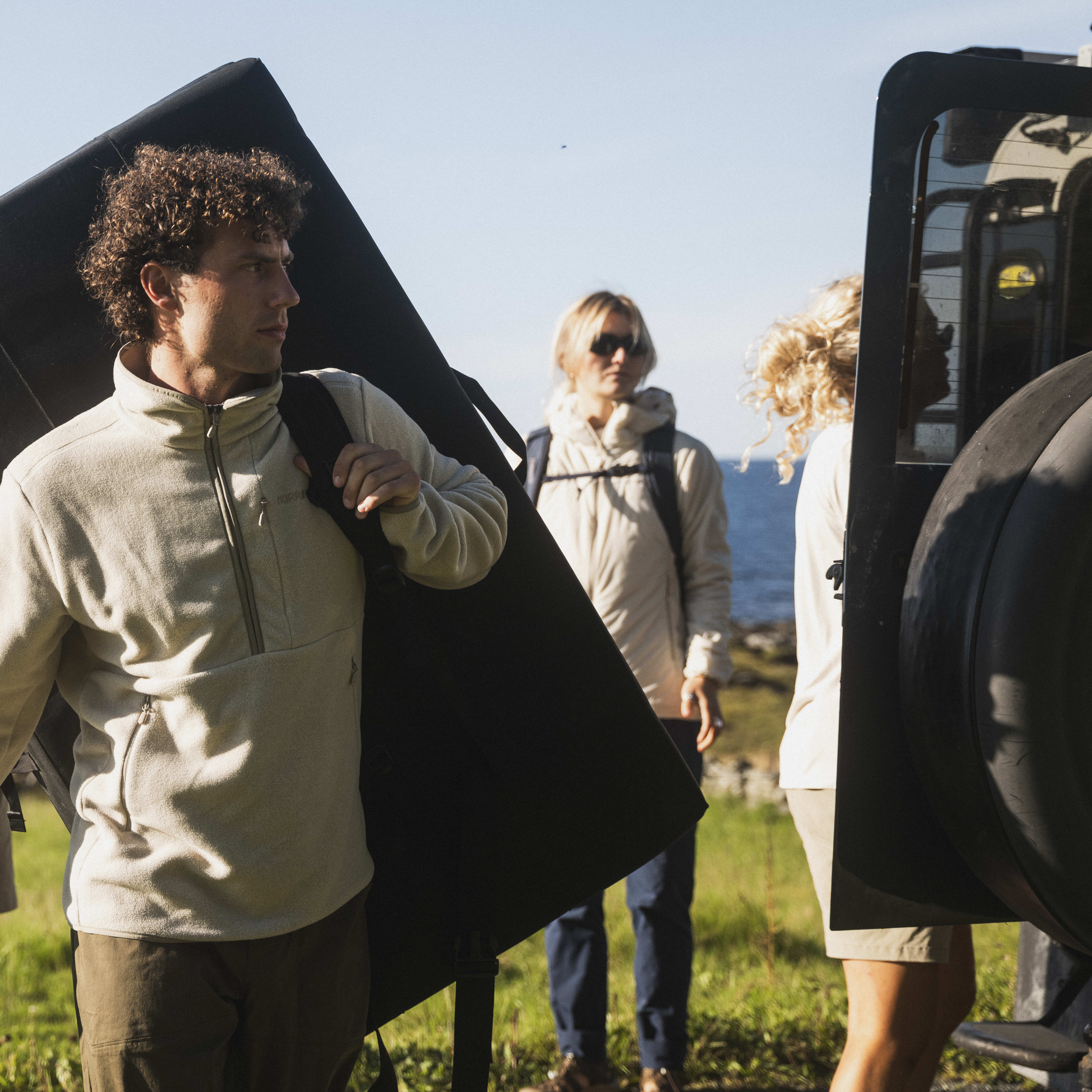 A man in a Norrøna fleece carries a crash pad with others near a car. They are outdoors, with a glimpse of the sea in the background.
