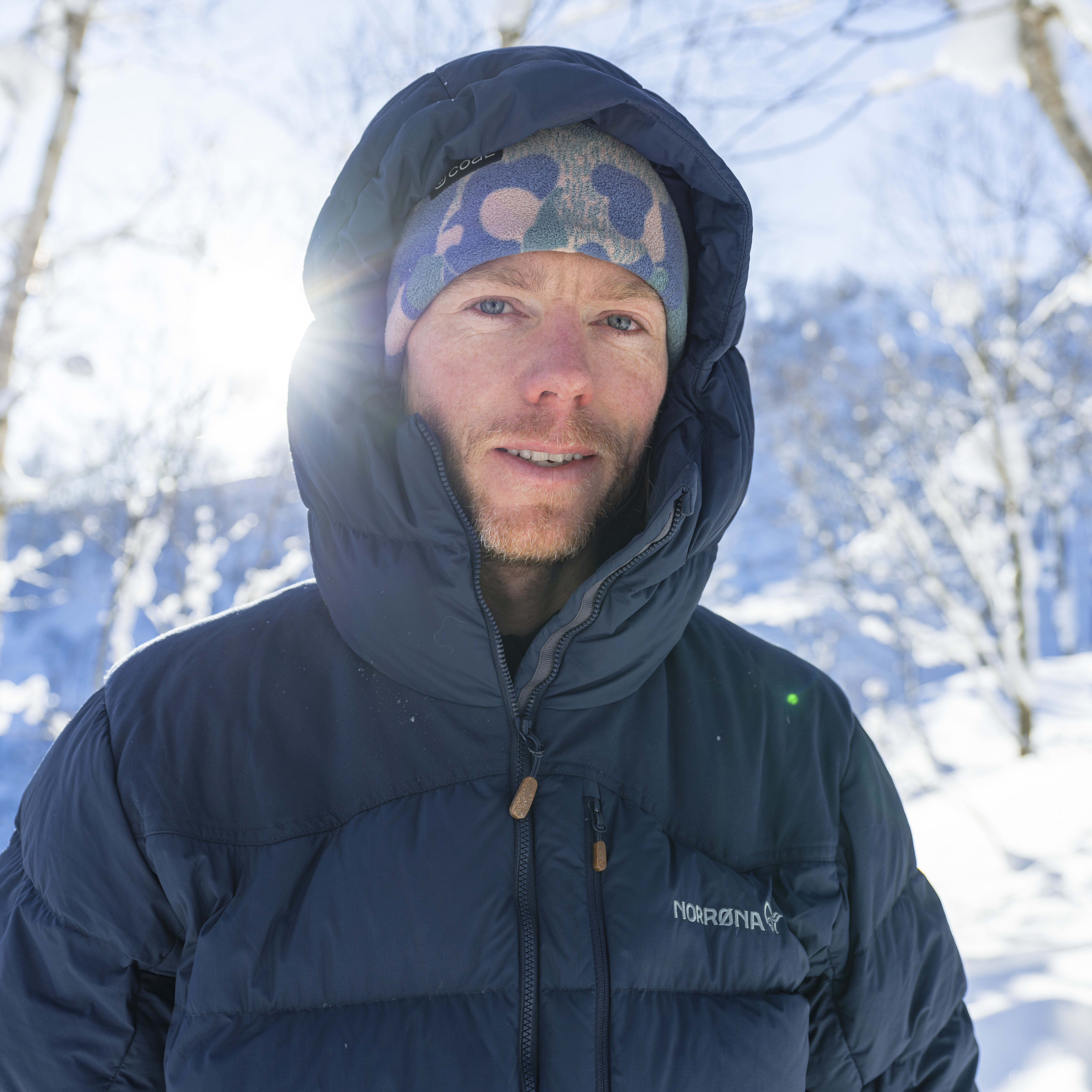 A man wearing a Norrøna jacket and a patterned hat stands in a snowy forest. The sun shines in the background. 
