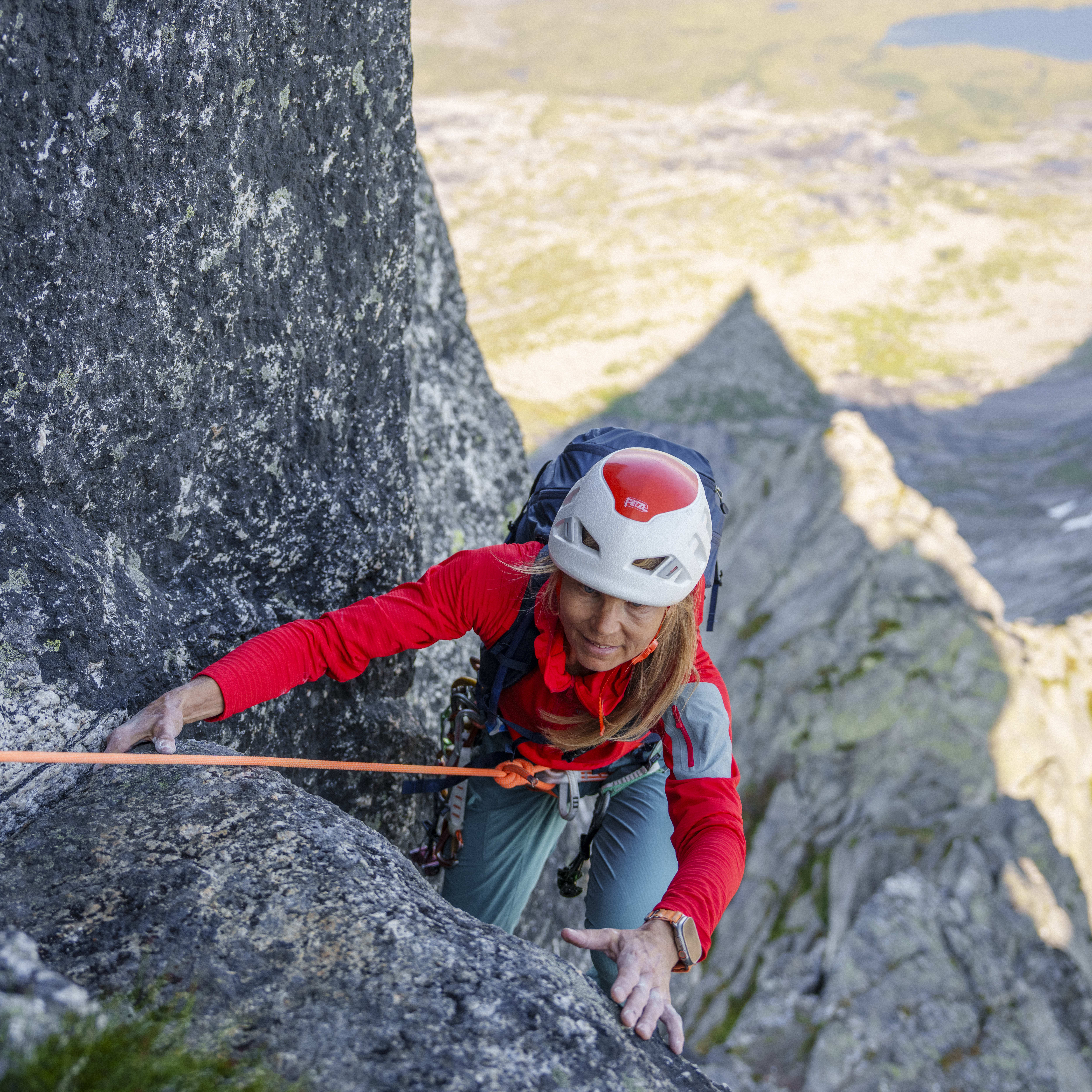 A woman rock climbs, secured by rope. She wears a helmet, pack, red Norrøna shirt, and blue pants; a valley is visible in the background.