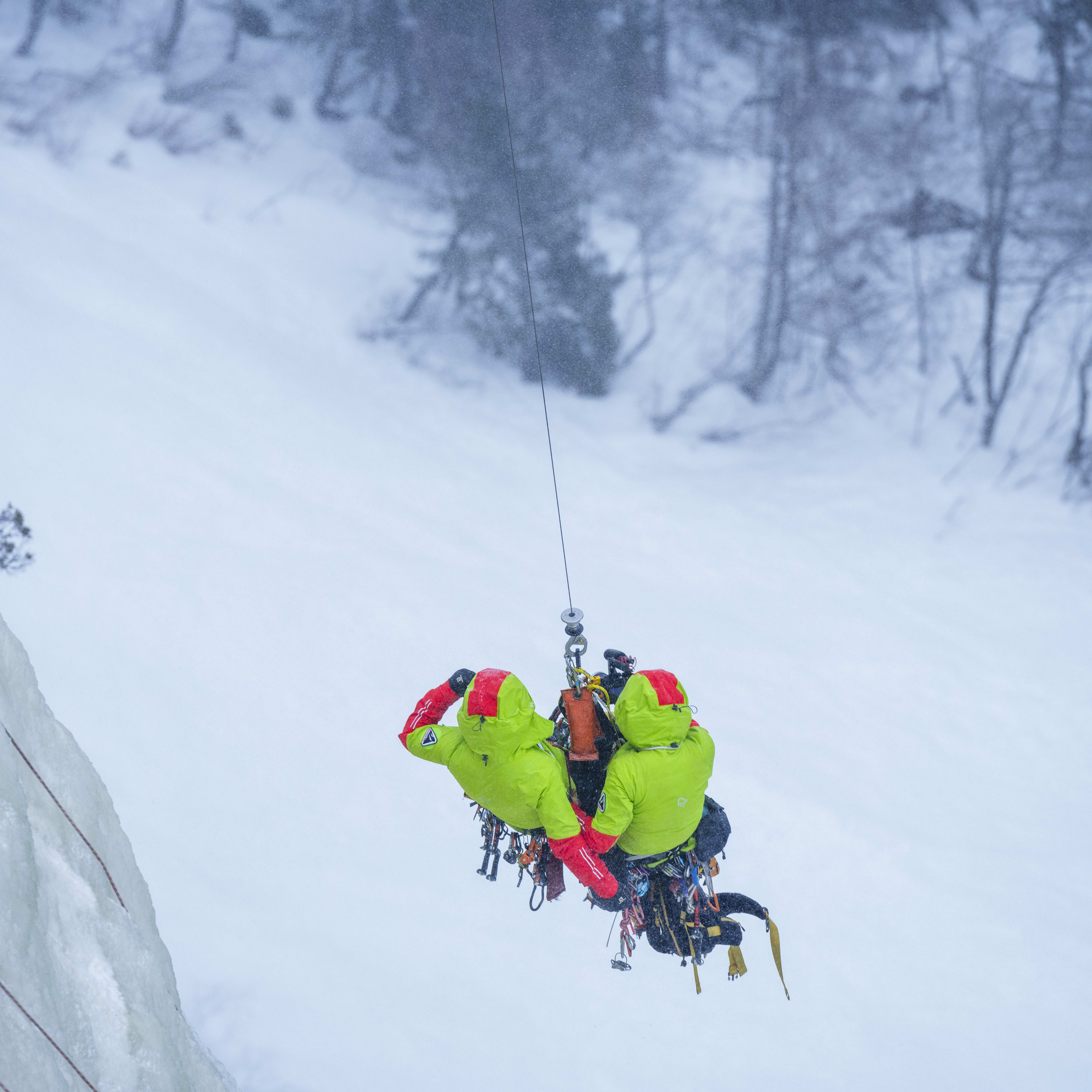 Two ice climbers rappelling down a snowy cliff face. They are wearing Norrøna jackets.  
