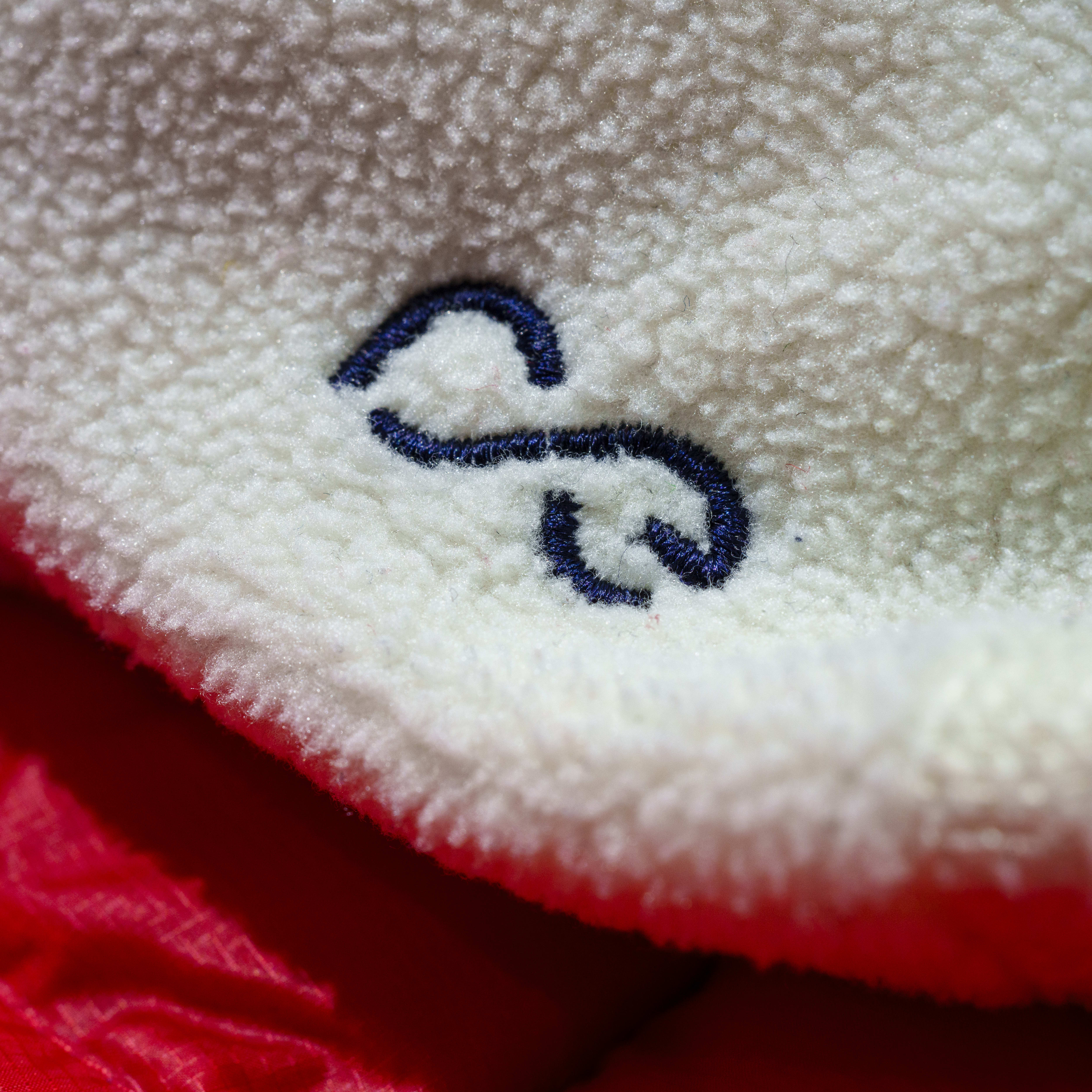 Close-up of a white fuzzy fabric with a blue infinity symbol embroidered on it. The fabric is folded over a red fabric.
