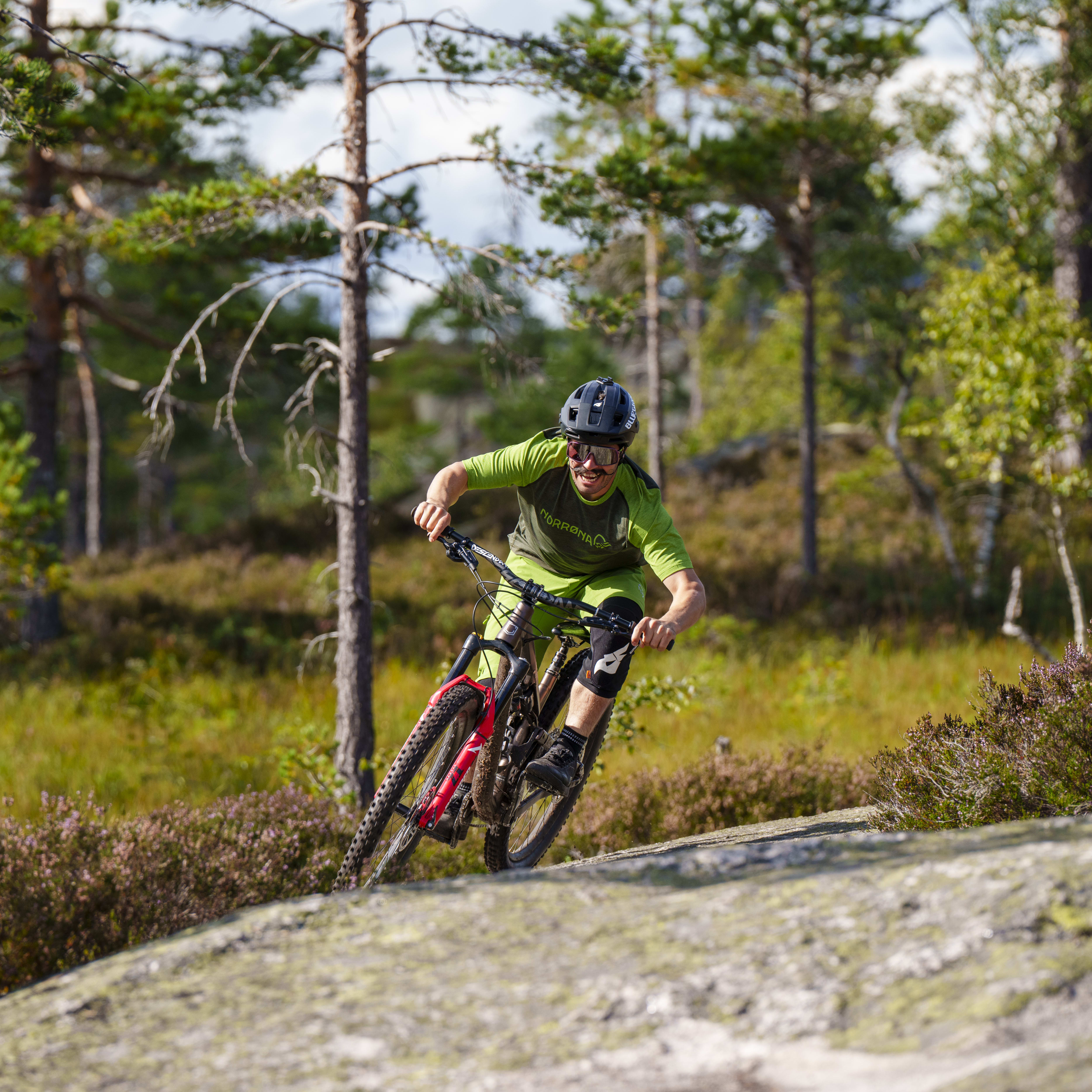 A man wearing a green Norrøna shirt, black pants, and a helmet,  rides a black and red mountain bike through a forest. The bike is in motion, with the front wheel lifted slightly off the ground.  The man is smiling and appears to be enjoying the ride.