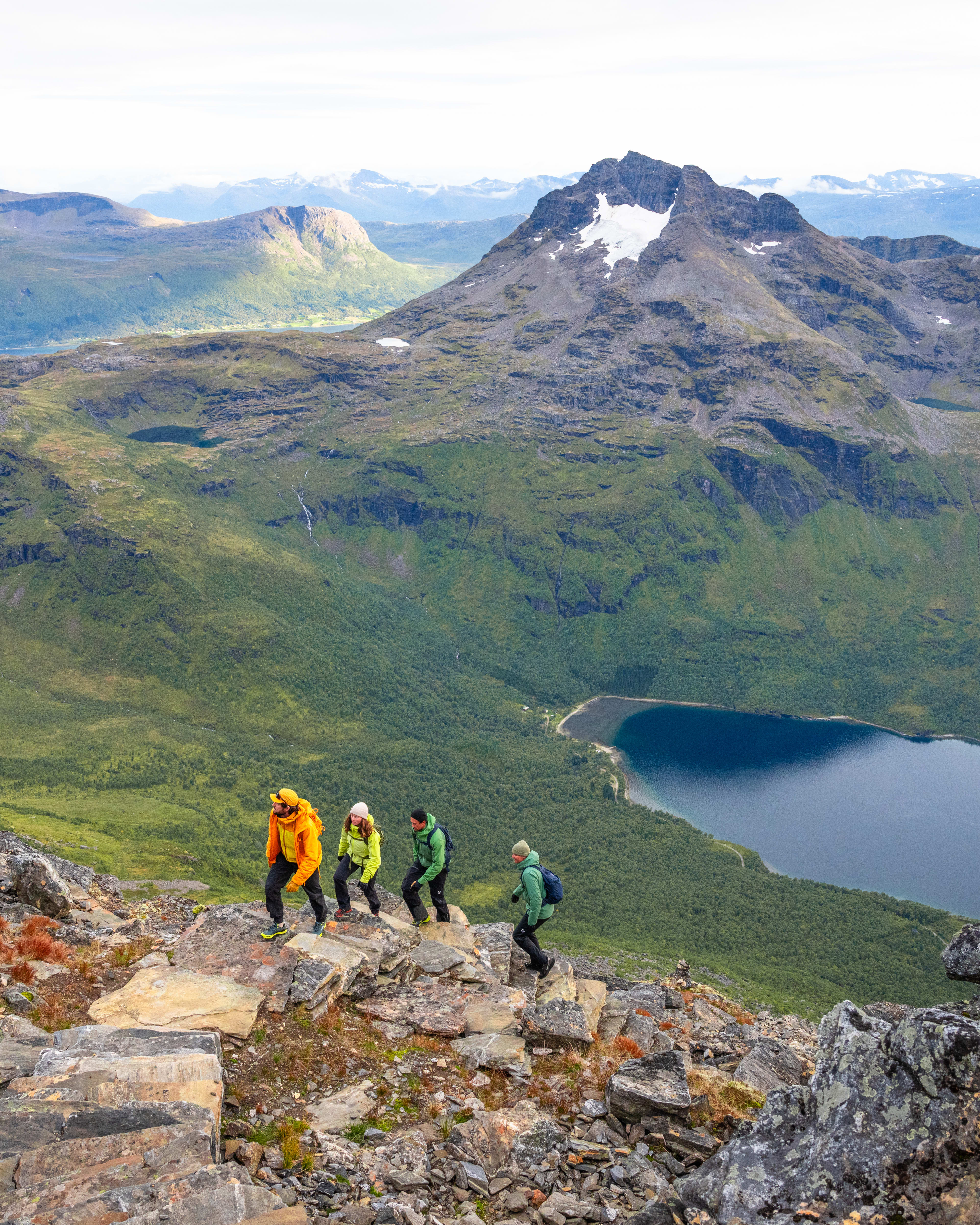 Four hikers in Norrøna gear walk along a rocky mountain ridge overlooking a deep blue lake.
