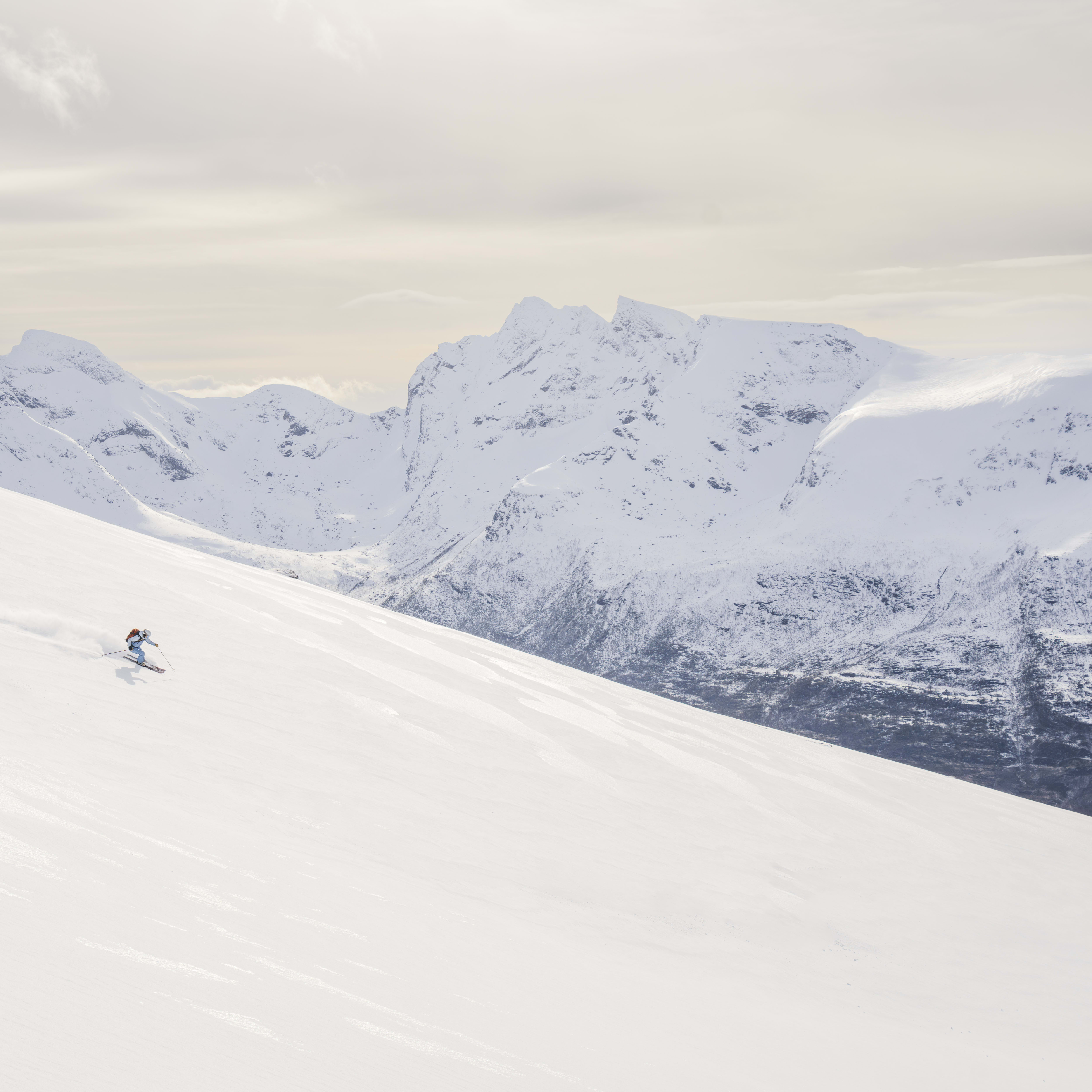 A skier wearing Norrøna apparel carves a turn on a vast, snow-covered mountain with jagged, snowy peaks in the background under a cloudy sky.