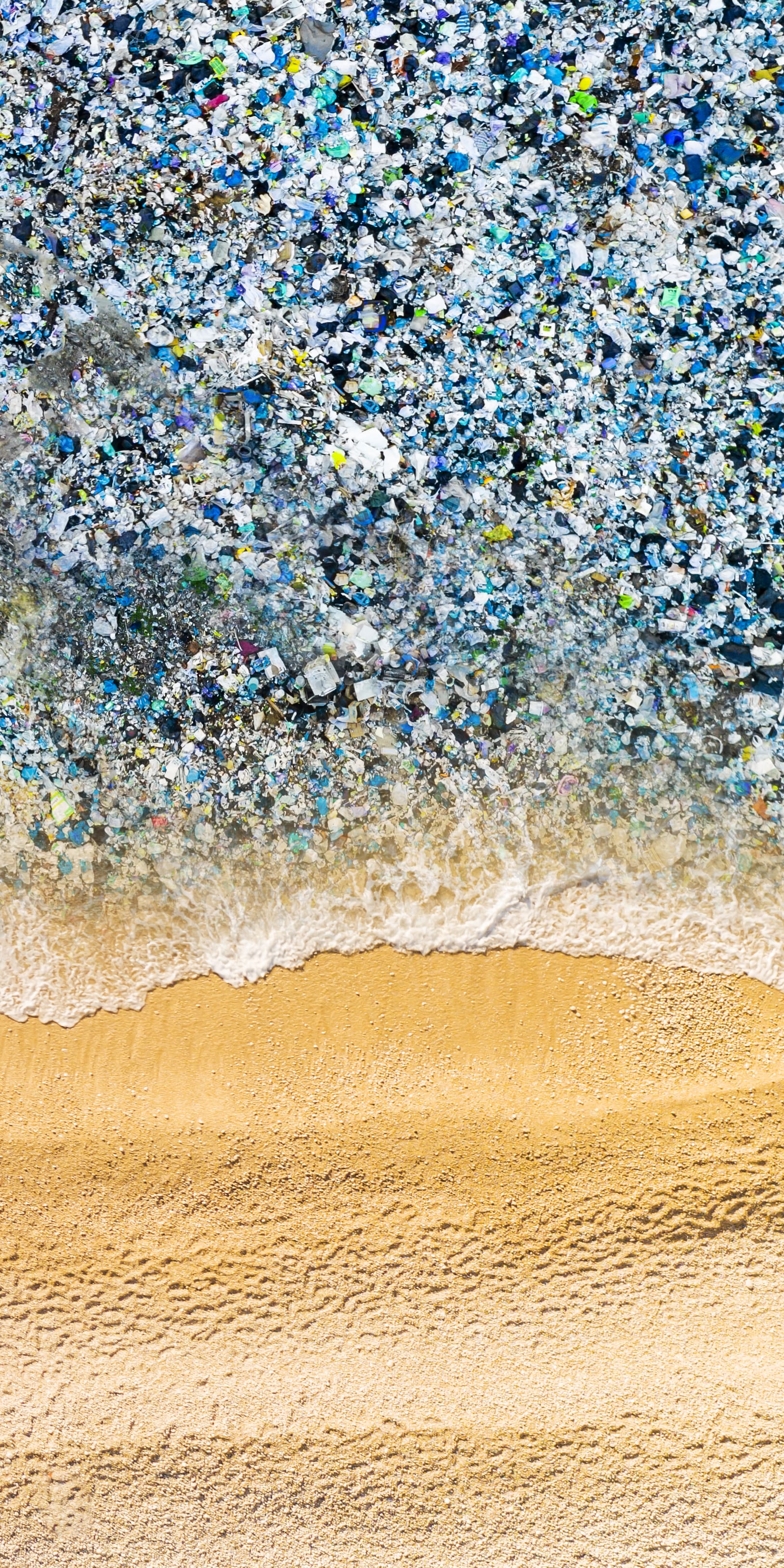 Aerial view of a beach polluted with plastic waste and a wave rolling onto the shore. 

