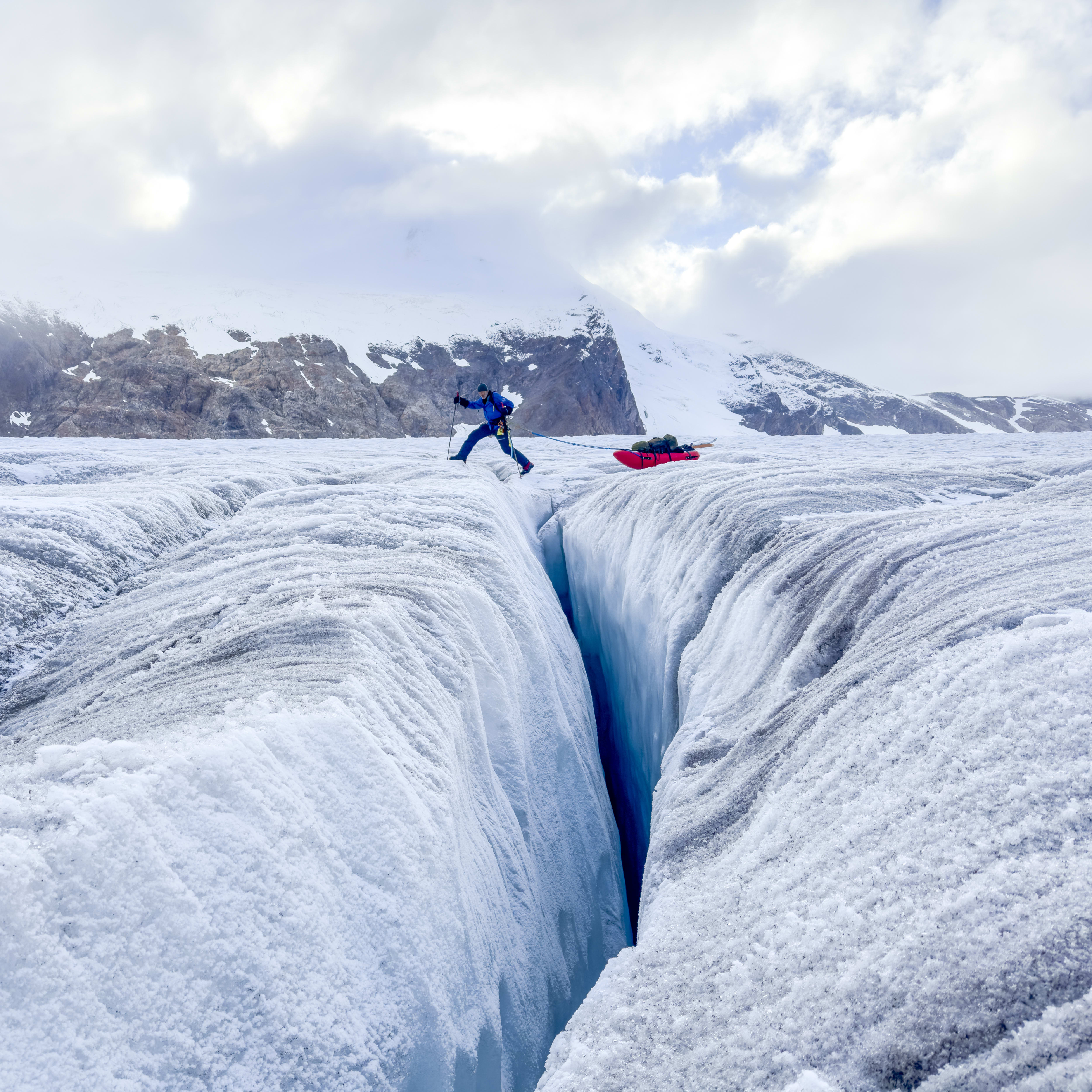 A person in Norrøna clothing leaps over a crevasse on a glacier, with a red sled and mountains in the background.