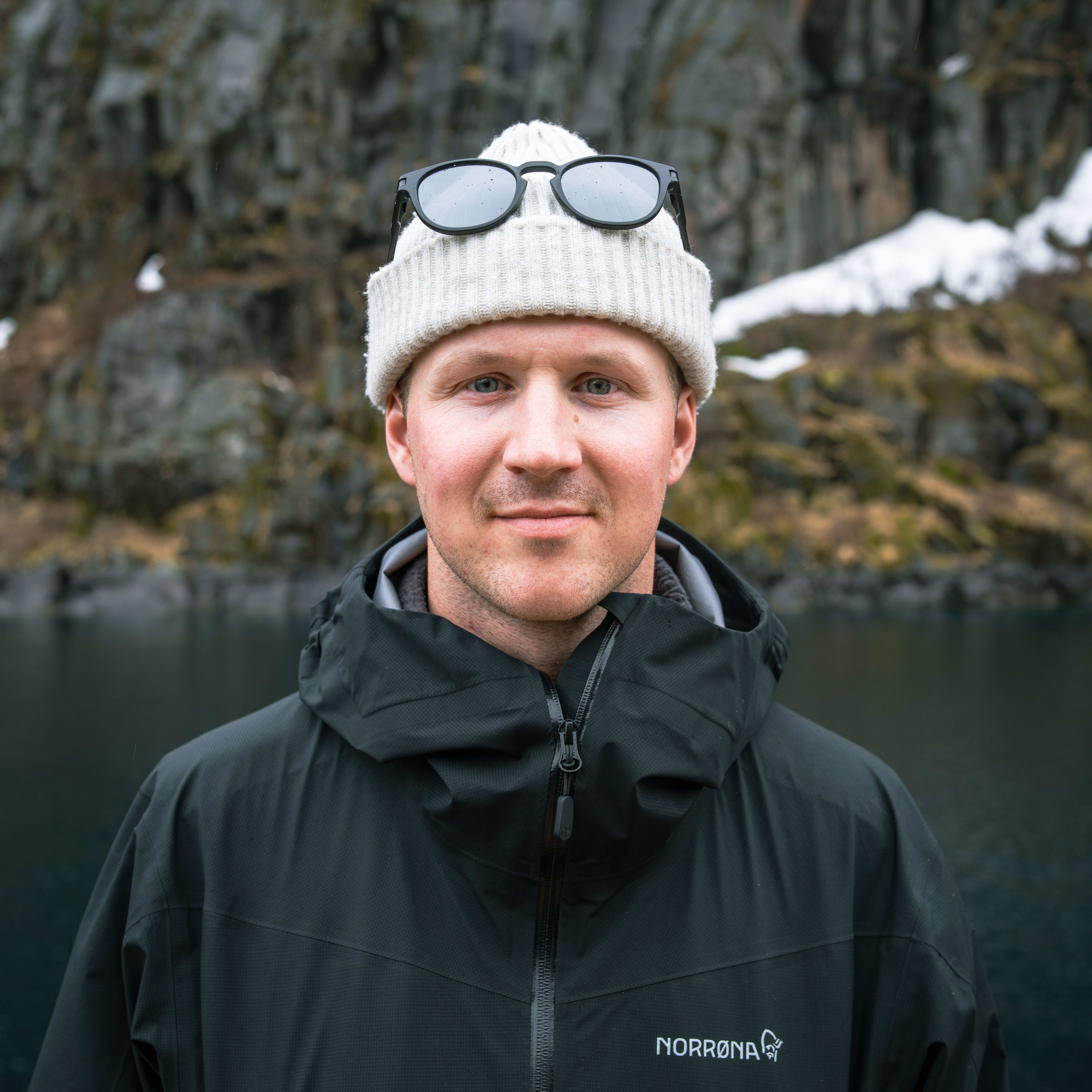 A man wearing a white knit hat, sunglasses and a black Norrøna jacket, smiles in front of a lake and rocky hillside.