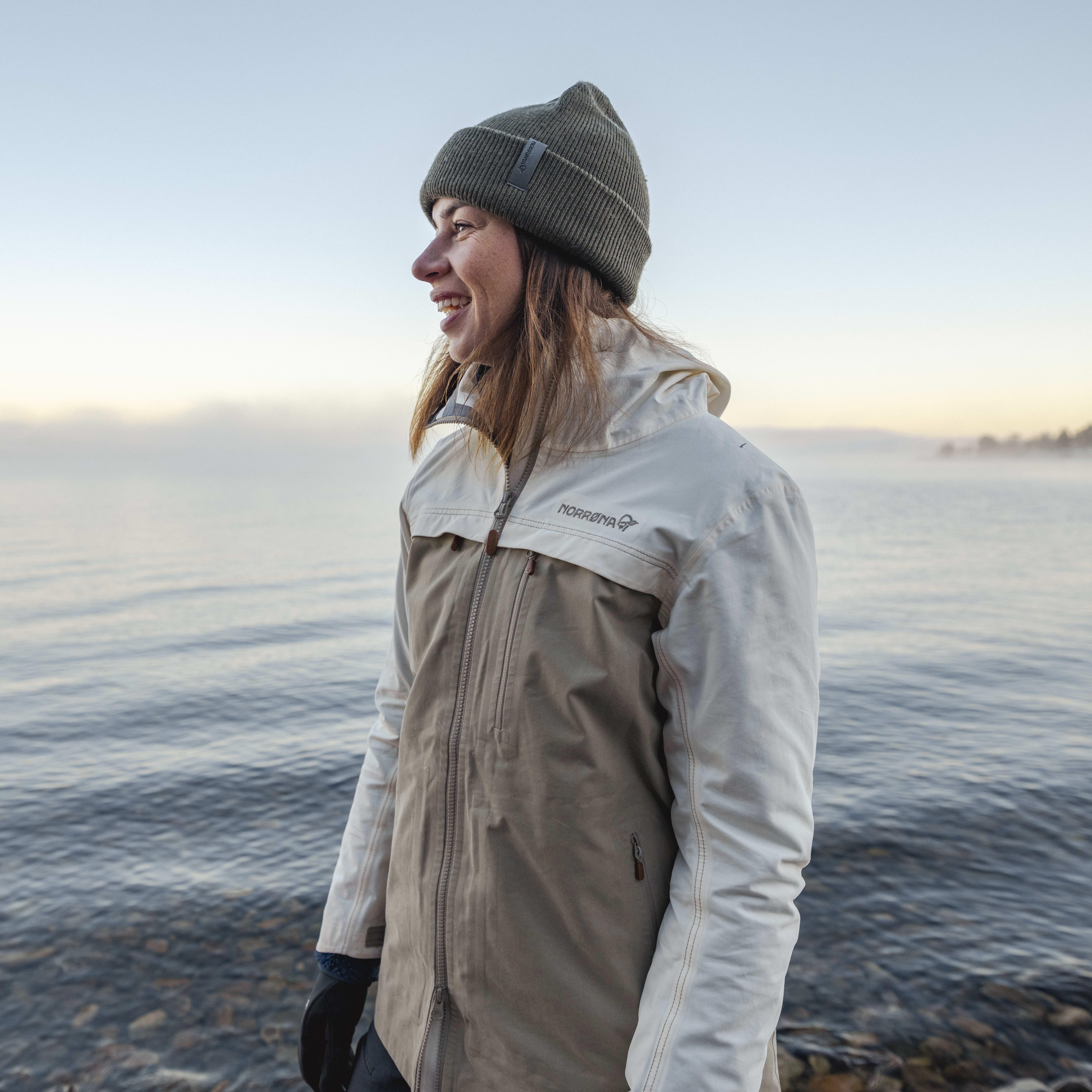 A woman in a Norrøna jacket and hat smiles at the camera while standing on a lakeshore. 
