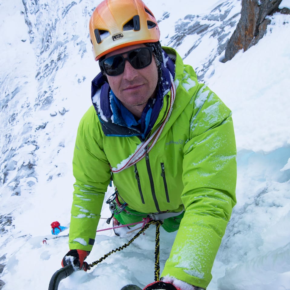 A man ice climbing, looking towards the camera. He wears a helmet, sunglasses, and a green Norrøna jacket. Ice axes are visible.
