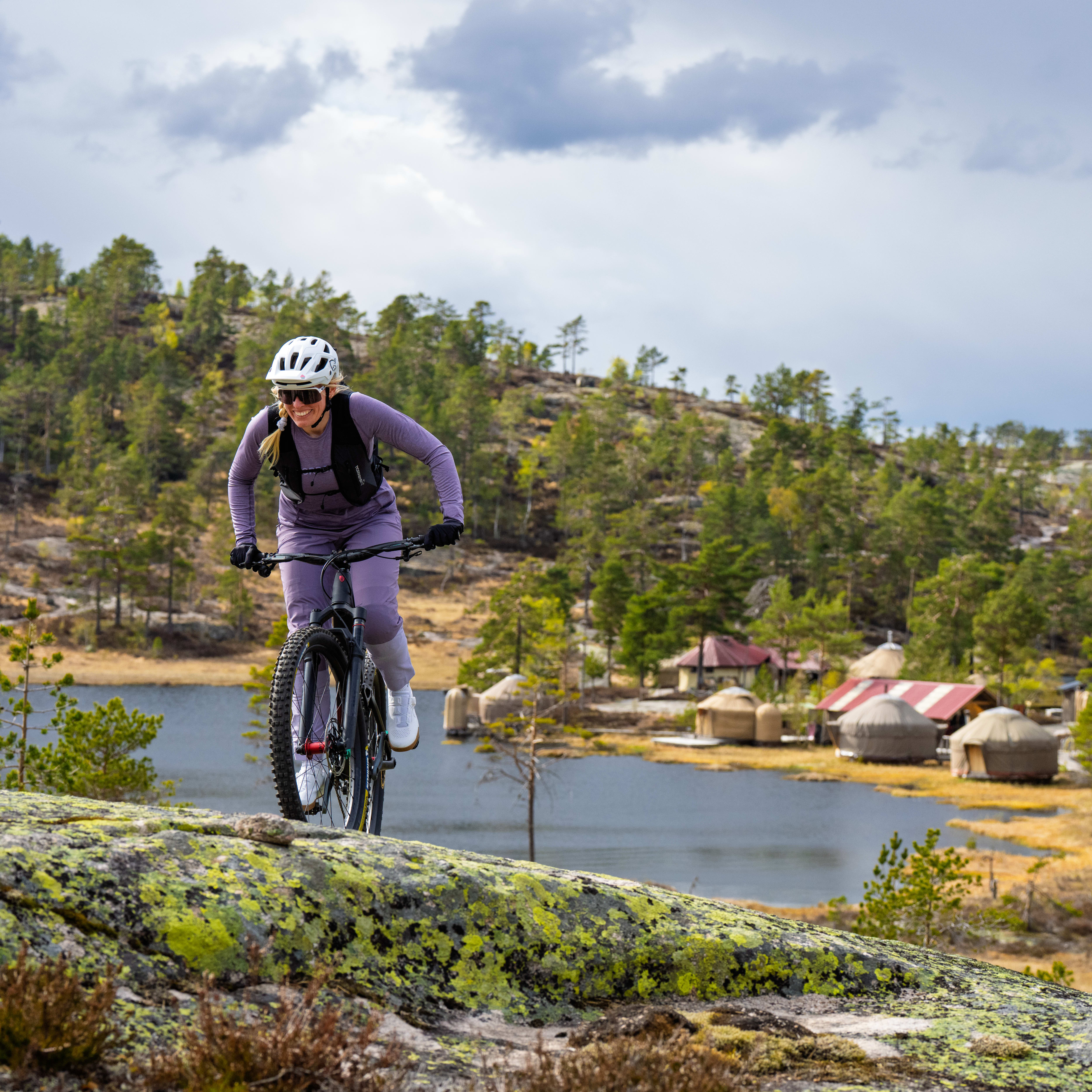 Woman in purple Norrøna gear mountain biking on a rocky ridge with a lake in the background.