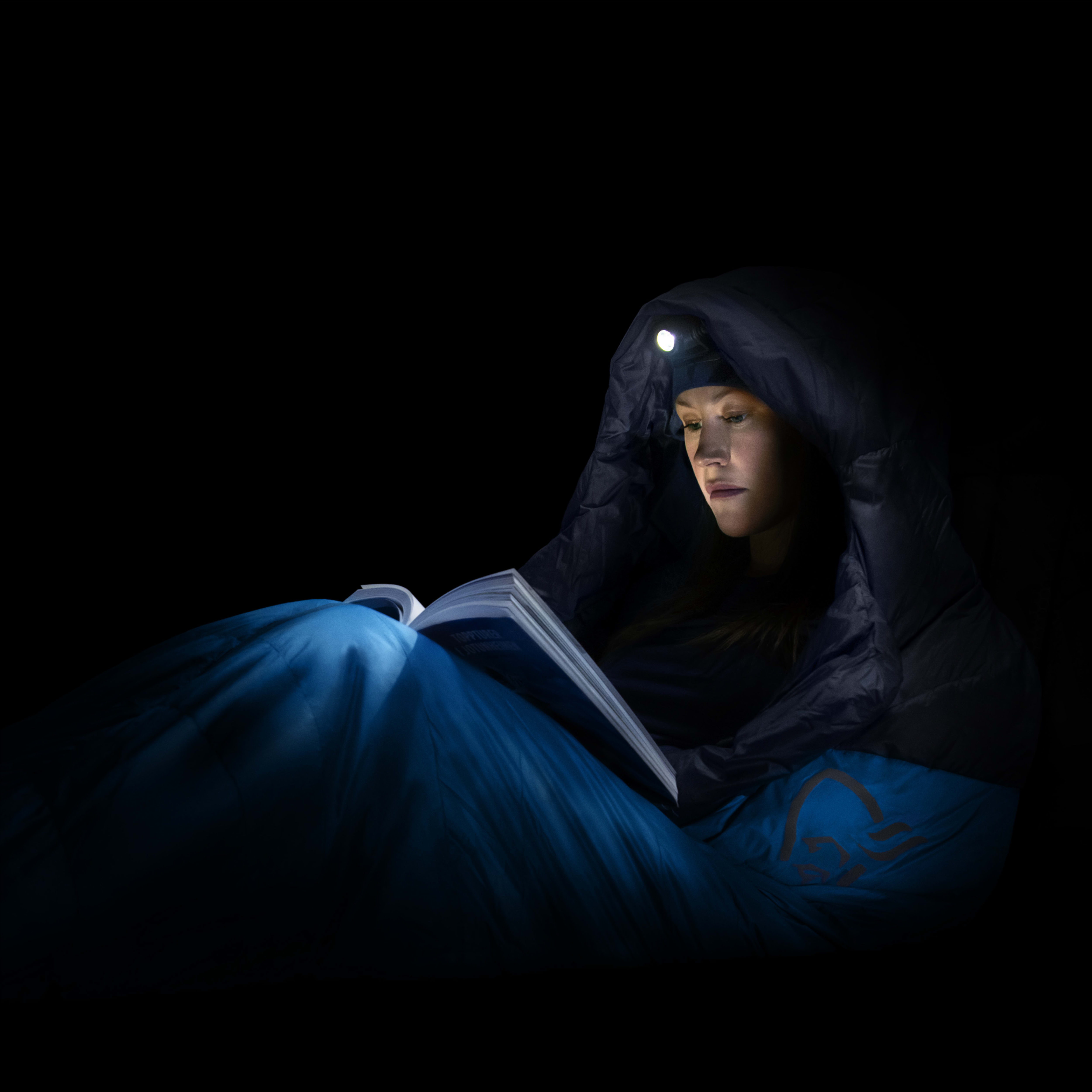 A person in a blue Norrøna sleeping bag with a headlamp is reading a book in the dark.