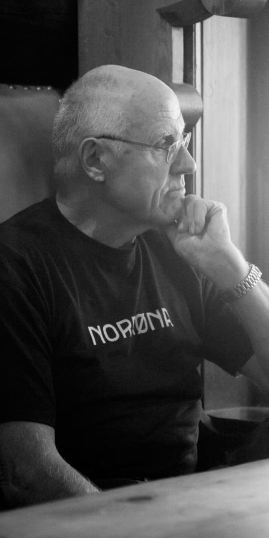 A man wearing a Norrøna shirt sits at a table, looking thoughtfully out a window. 
