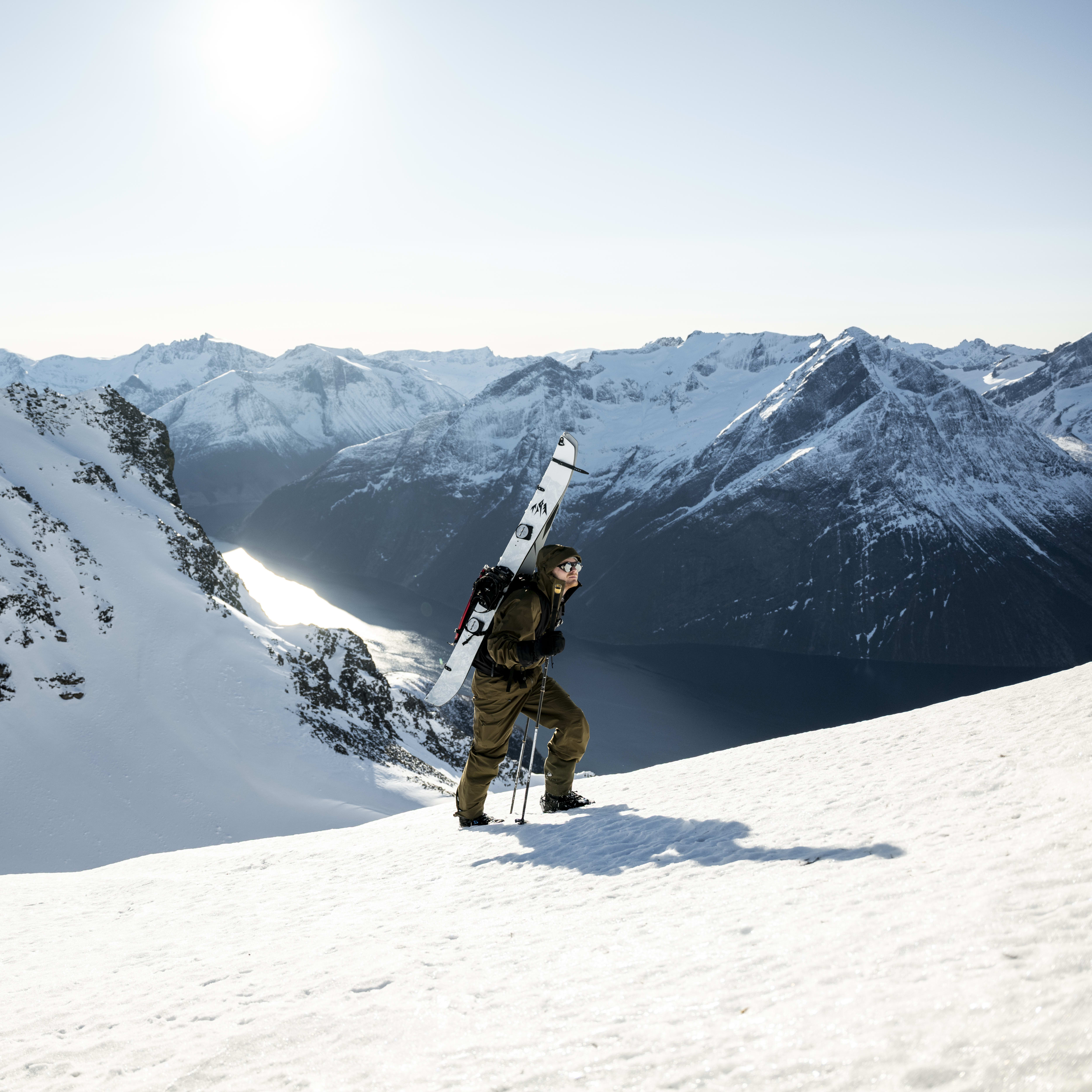 A man in Norrøna pants hikes uphill with skis and poles against a snowy mountain and valley backdrop.