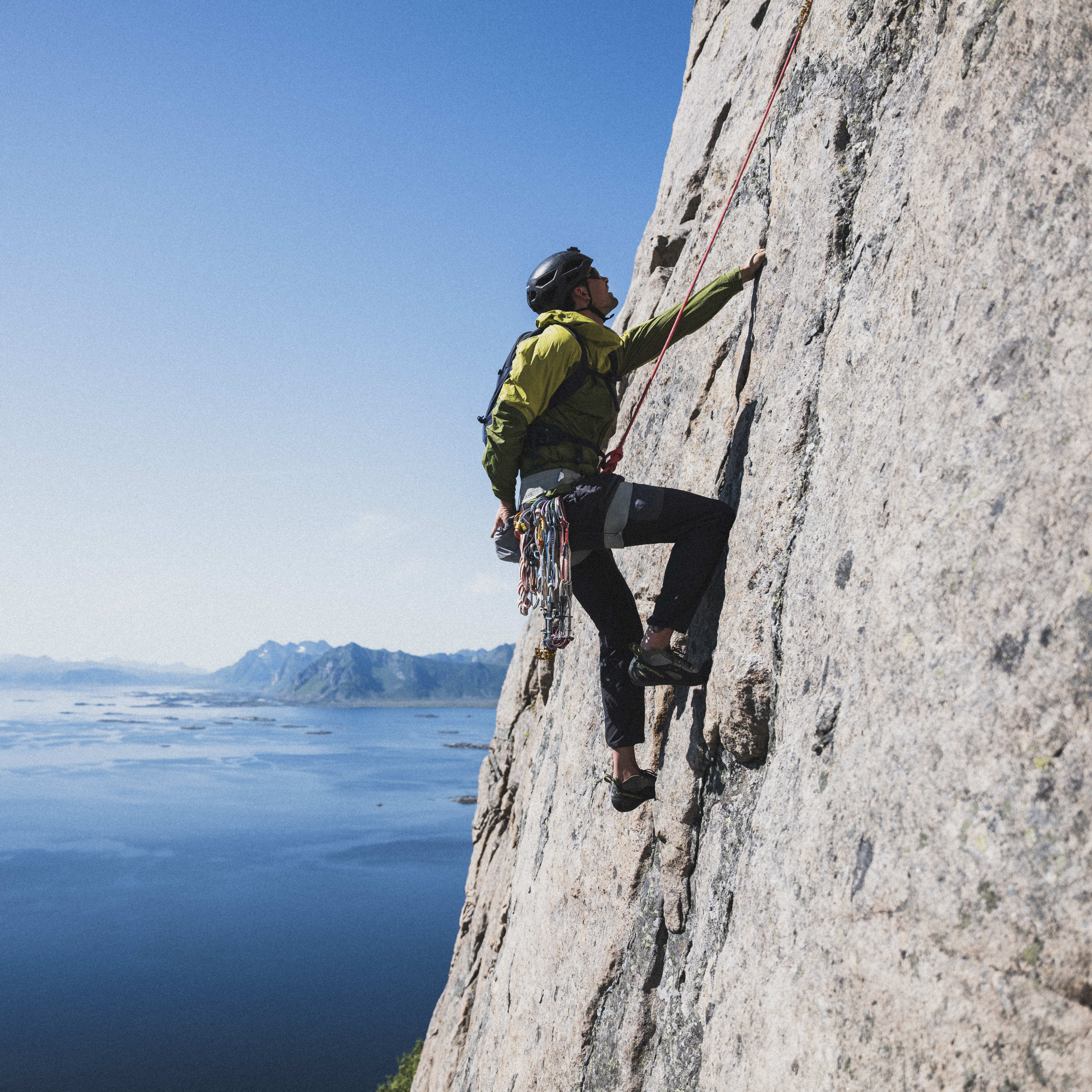 A climber in a Norrøna jacket scales a cliff face, secured by a rope. A blue sea with distant islands is in the background under a clear sky.