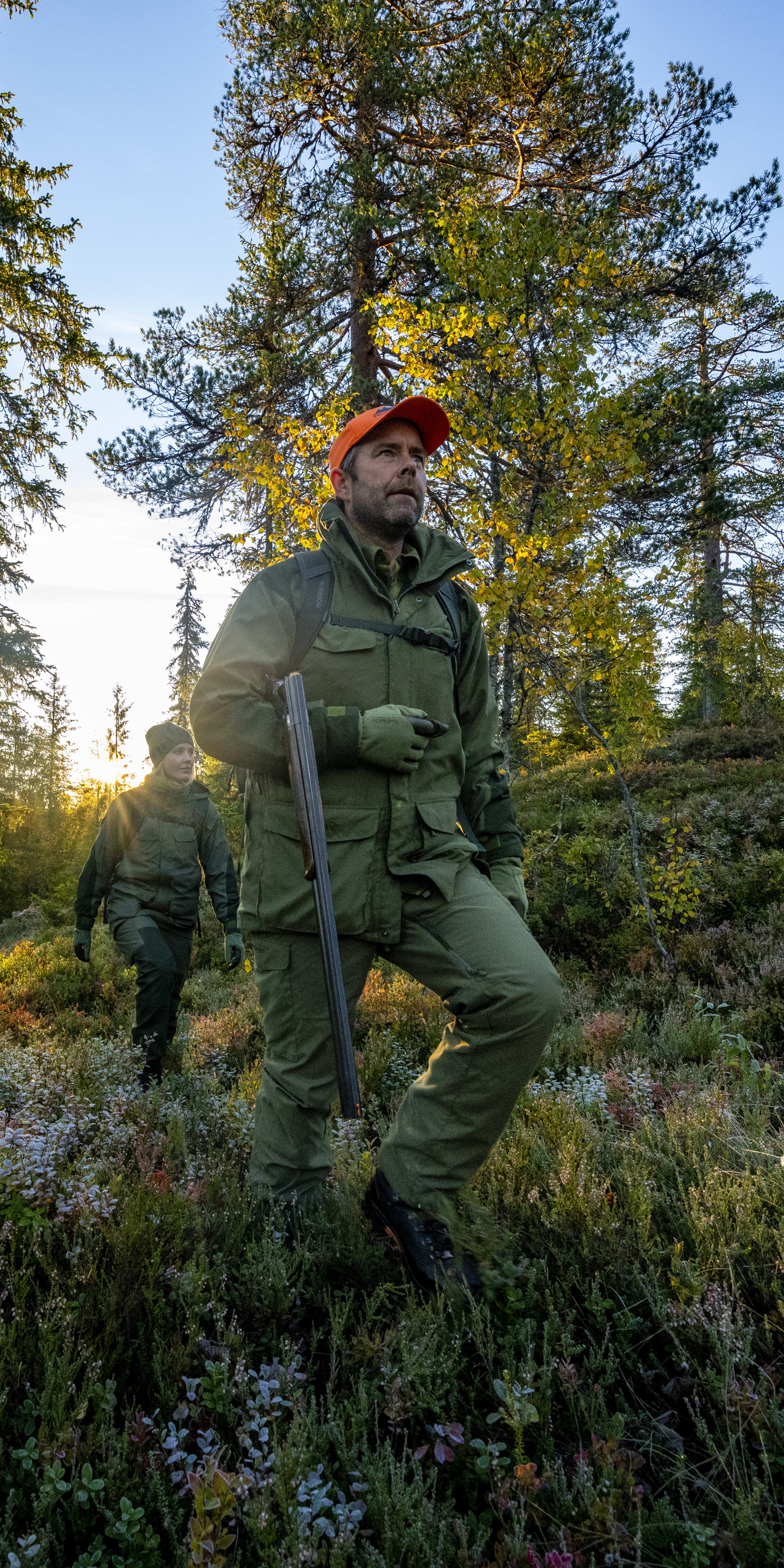 Two people wearing Norrøna clothing walk through a forest with a shotgun.  The sun shines through the trees. 
