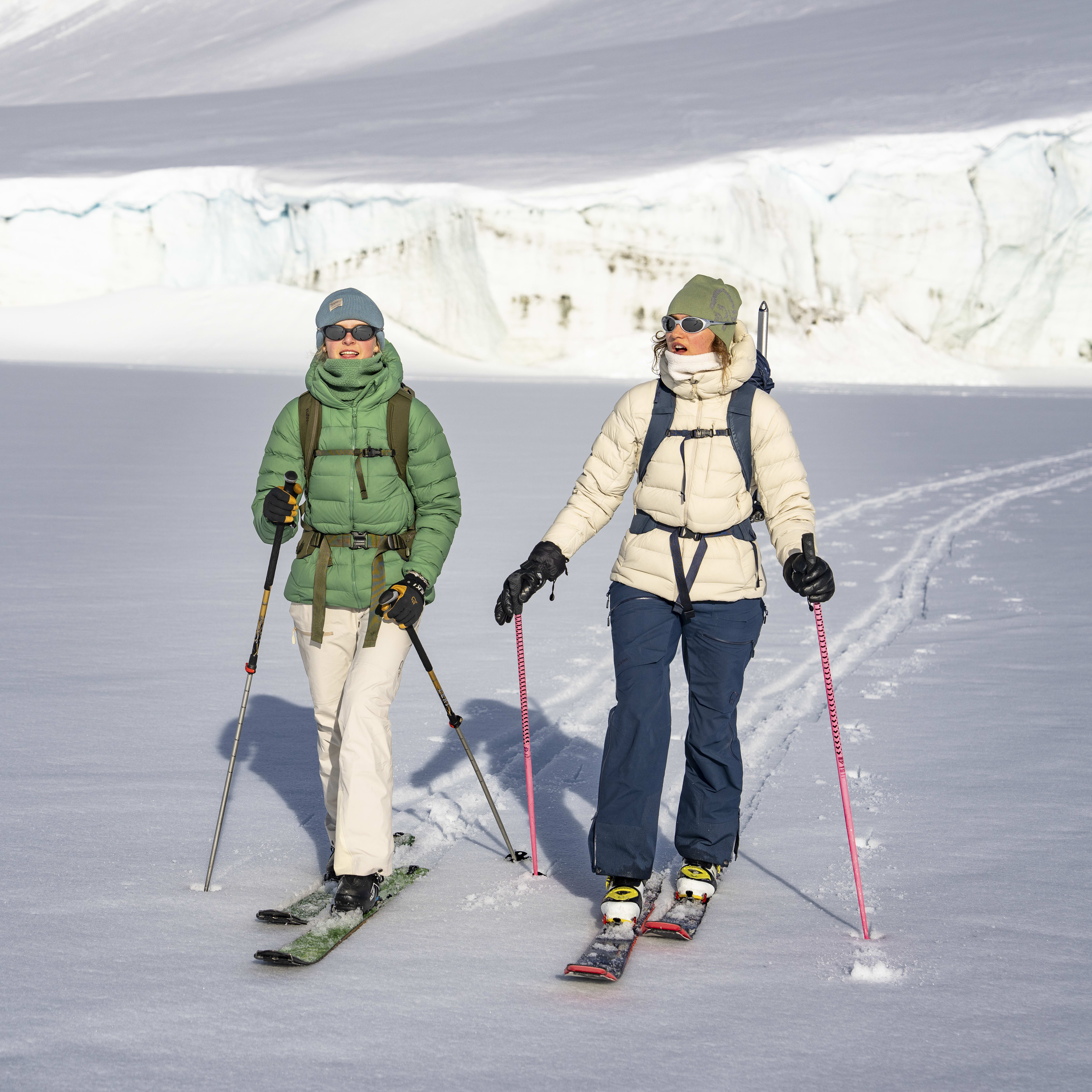 Two people in Norrøna clothing ski across a snowy landscape. They are wearing sunglasses and are smiling. 
