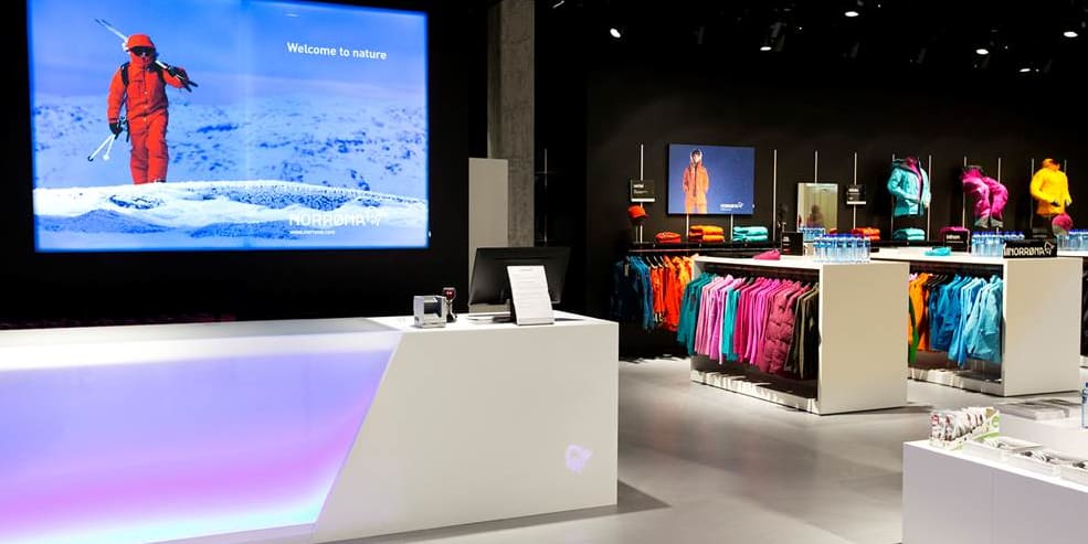 Interior of a Norrøna store featuring a modern illuminated counter and racks of colorful outdoor apparel.