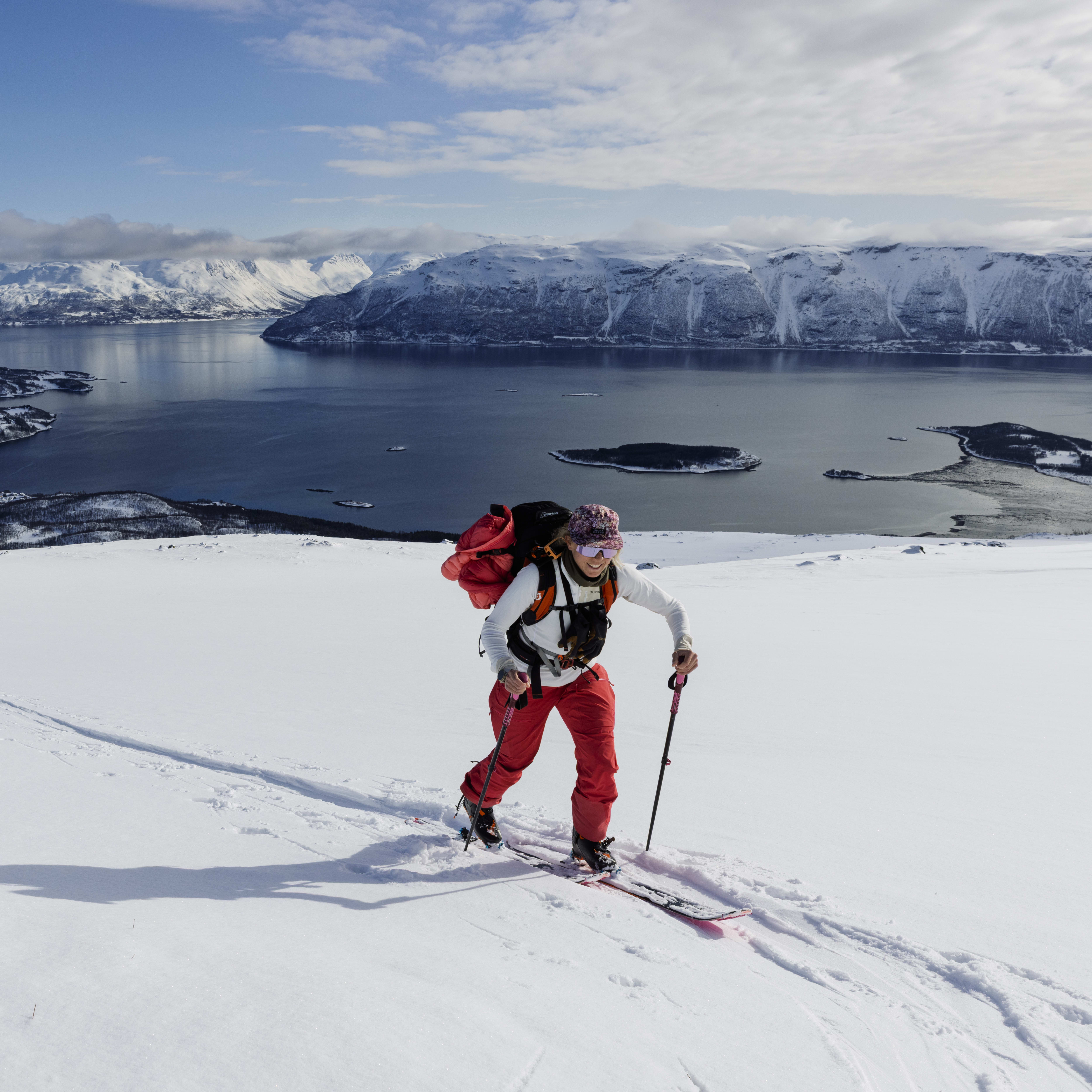 A person ski touring uphill in Norway's snowy landscape. The individual wears red Norrøna pants and carries a backpack with skis.