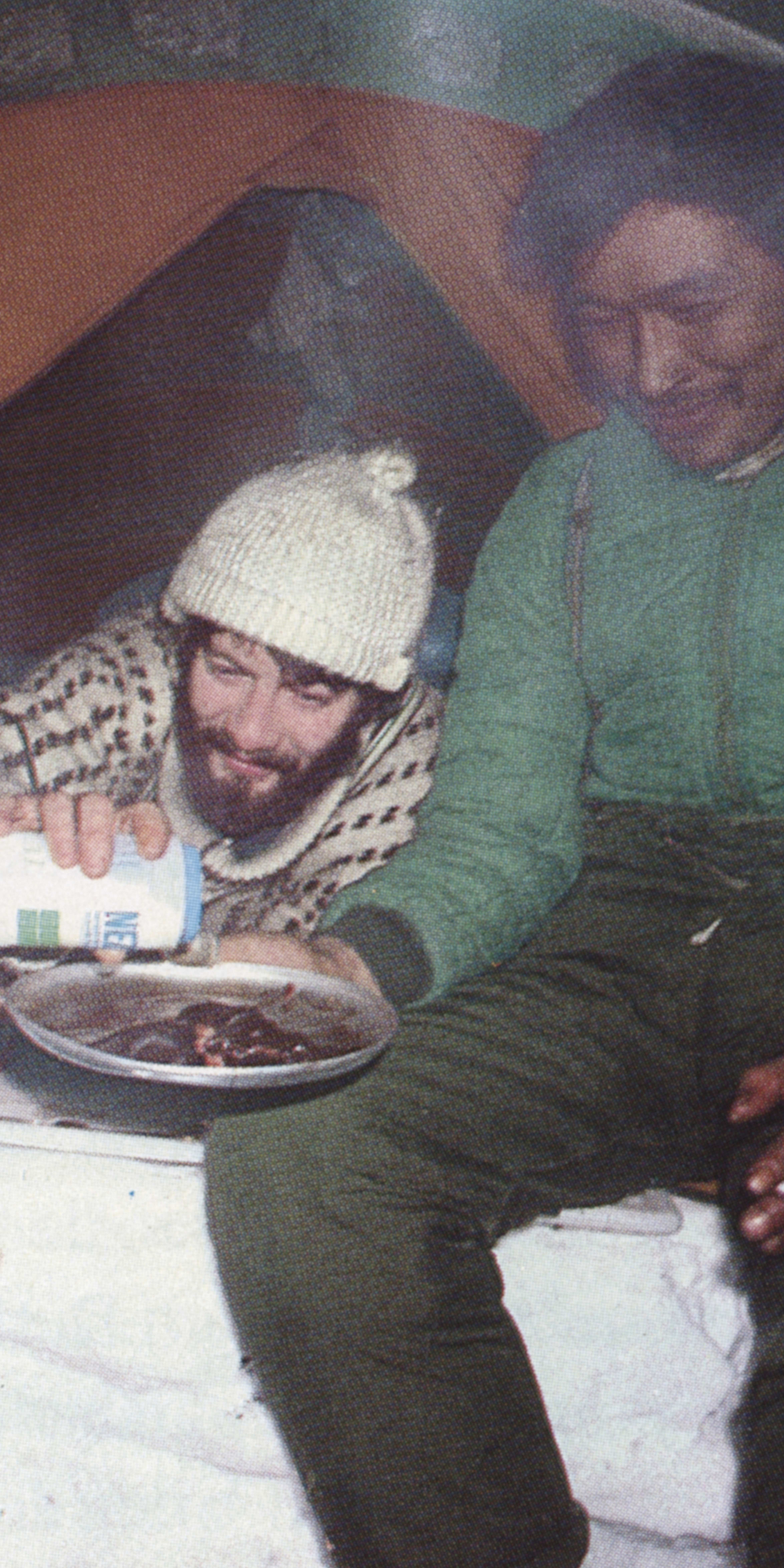 Two men are sitting in a tent. The man on the left is wearing a sweater and a white hat and is pouring something into a bowl. The man on the right is wearing a green jacket and has a cigarette in his hand. There is a cooking pot on the floor.
