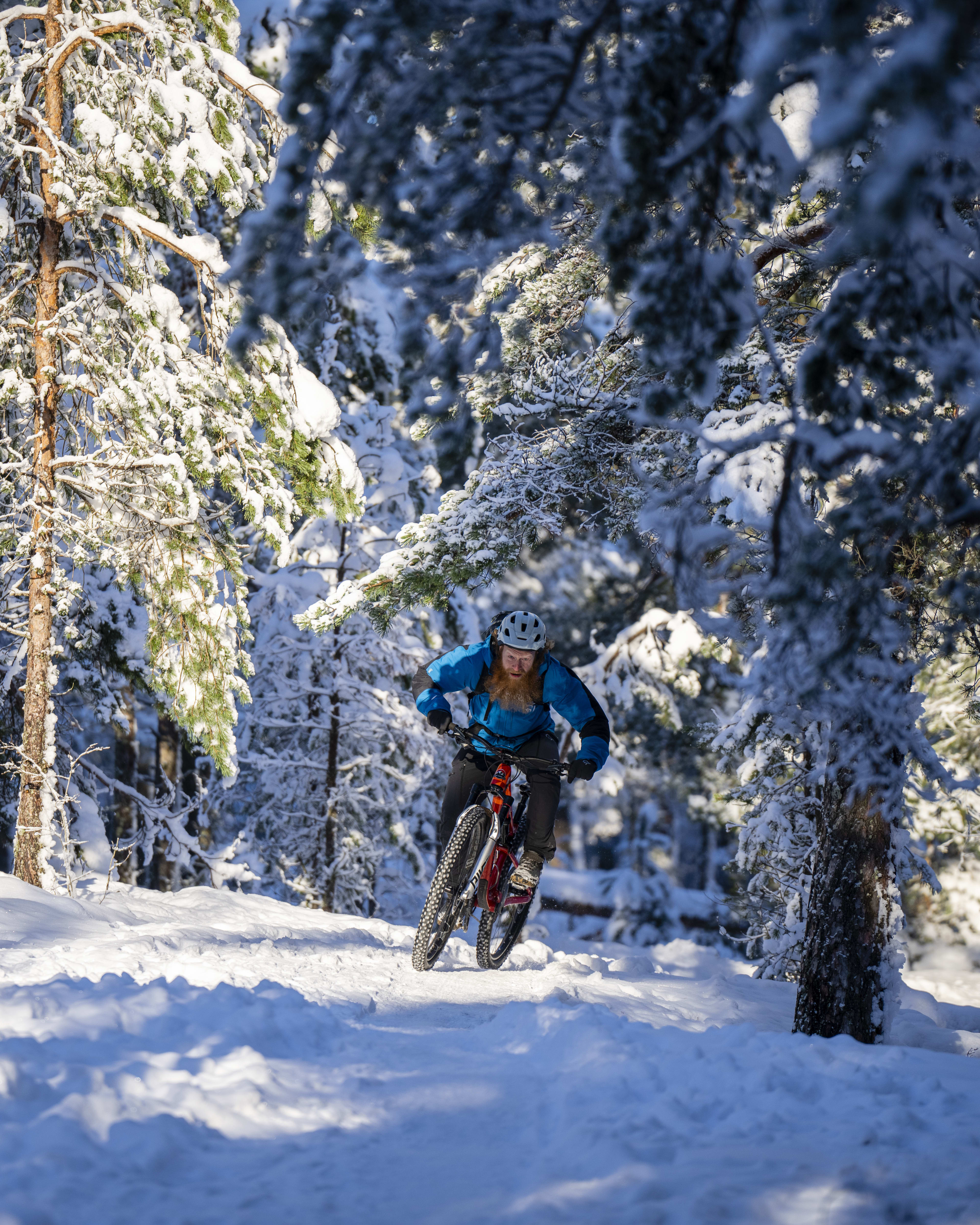 A man wearing a Norrøna jacket rides a mountain bike through a snowy forest.