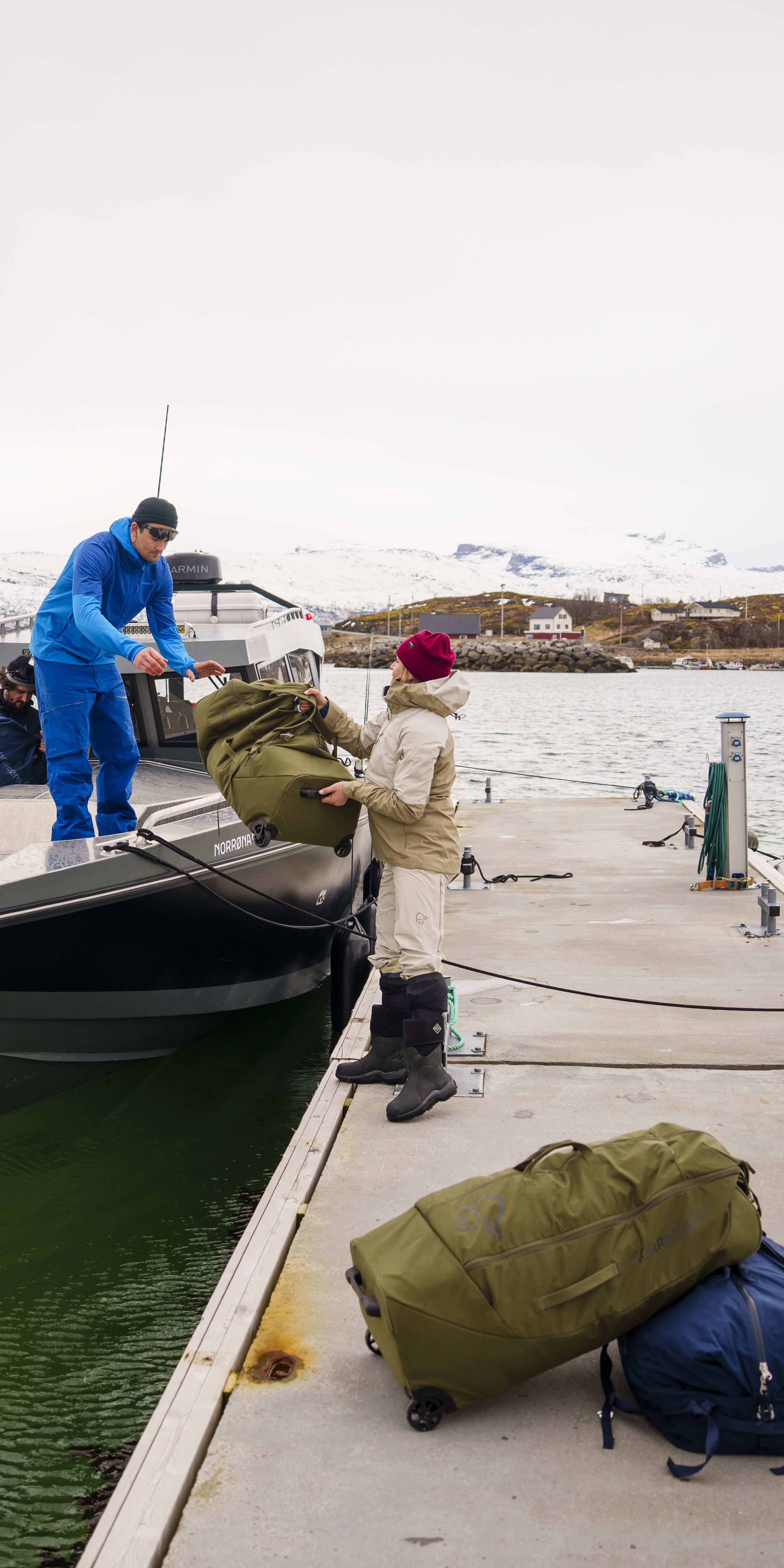Two people are on a dock, unloading a boat with luggage. One person is wearing a blue jacket from Norrøna and is on the boat. The other is wearing a tan jacket from Norrøna and is on the dock. Both are wearing Norrøna clothing. There are two pieces of luggage on the dock, both from Norrøna. The boat is docked by a wooden pier. There are mountains and water in the background.