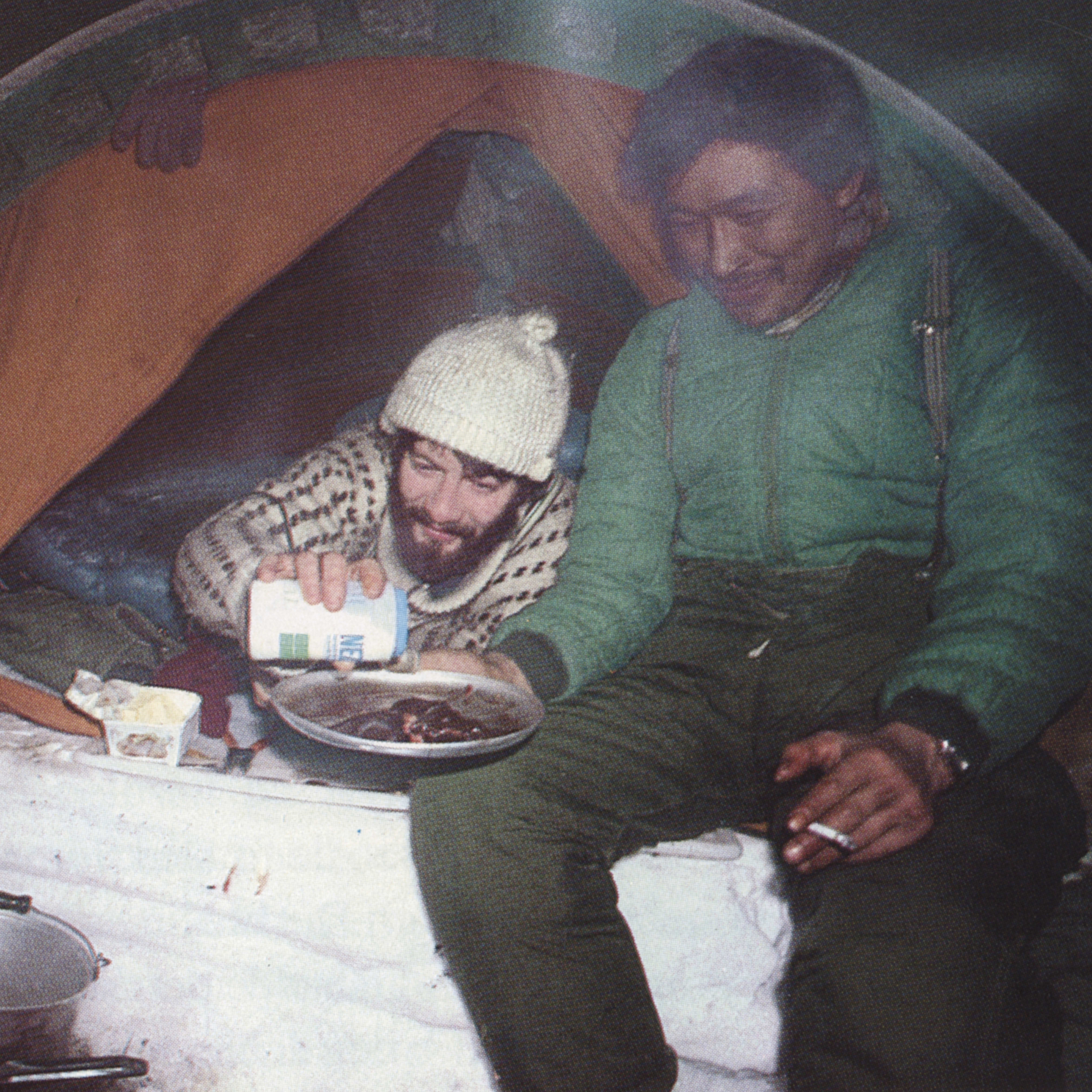 Two men are sitting in a tent. The man on the left is wearing a sweater and a white hat and is pouring something into a bowl. The man on the right is wearing a green jacket and has a cigarette in his hand. There is a cooking pot on the floor.
