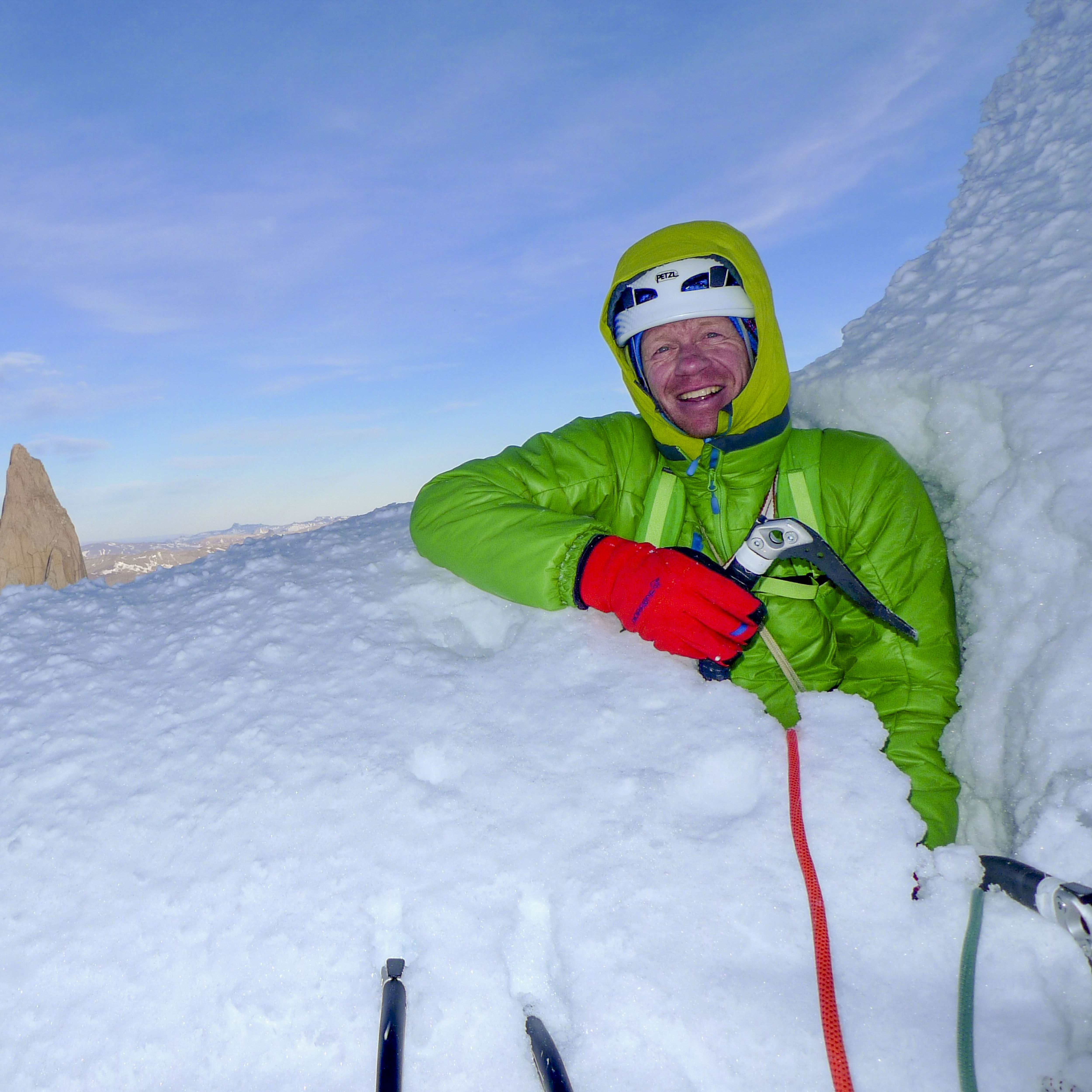 A man wearing a green jacket, red gloves, and a helmet is smiling at the camera. He is standing in the snow on a mountain peak, with a large rocky mountain in the background. Two ice axes and a red rope are in the snow around the man.