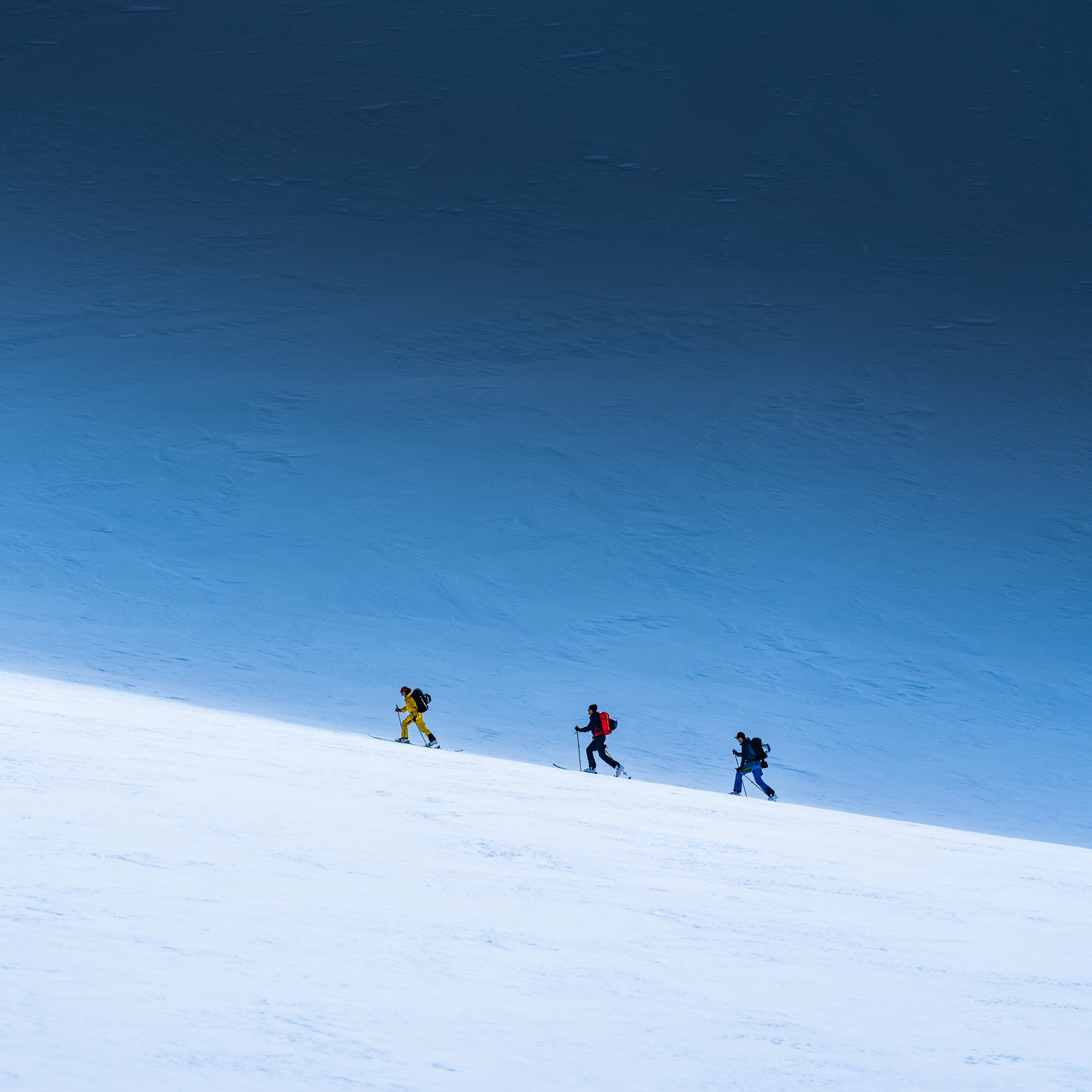 Three people in Norrøna gear ski uphill on a snowy mountain slope under a blue sky.