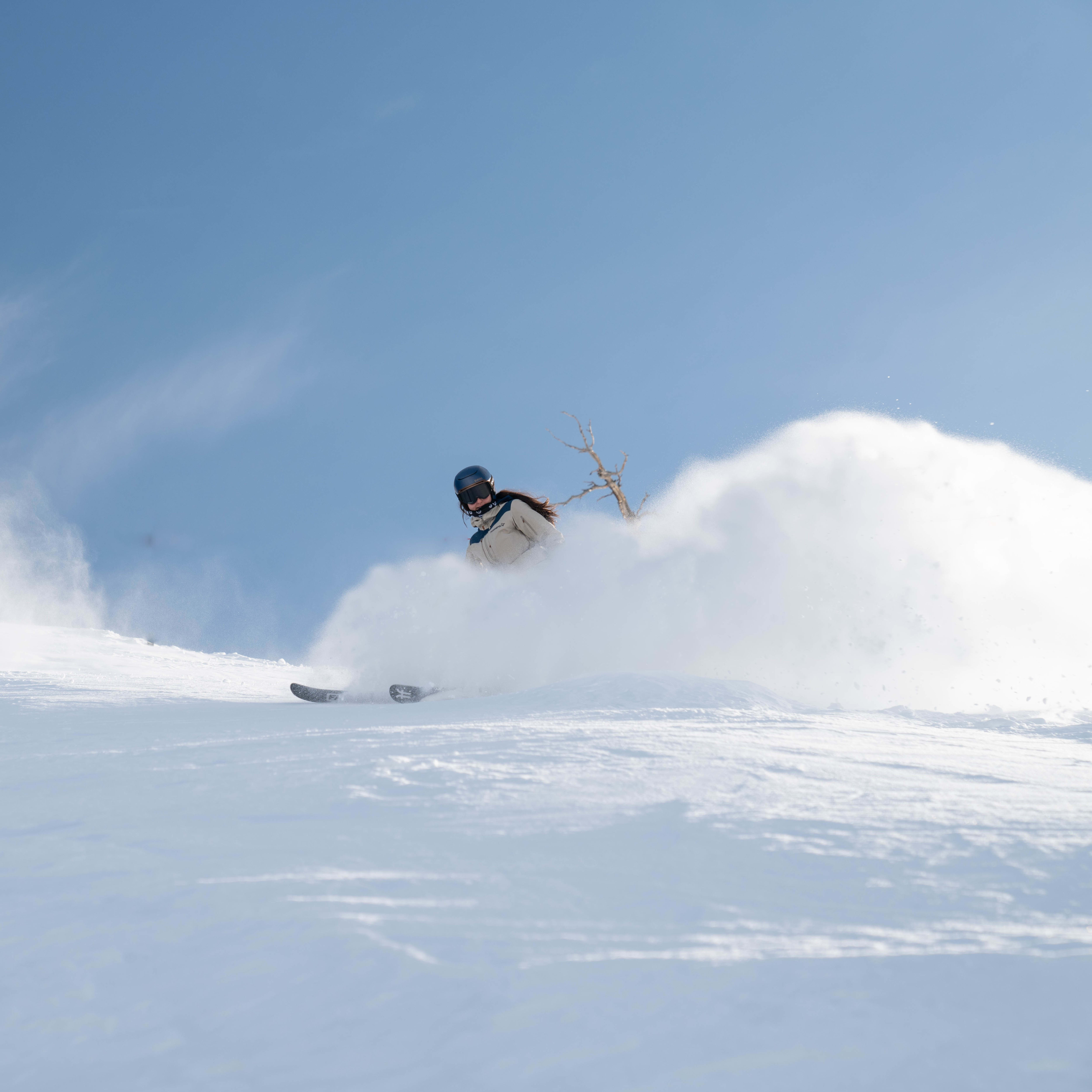 A skier in a Norrøna jacket carves through deep powder snow, kicking up a white spray against a clear blue sky.