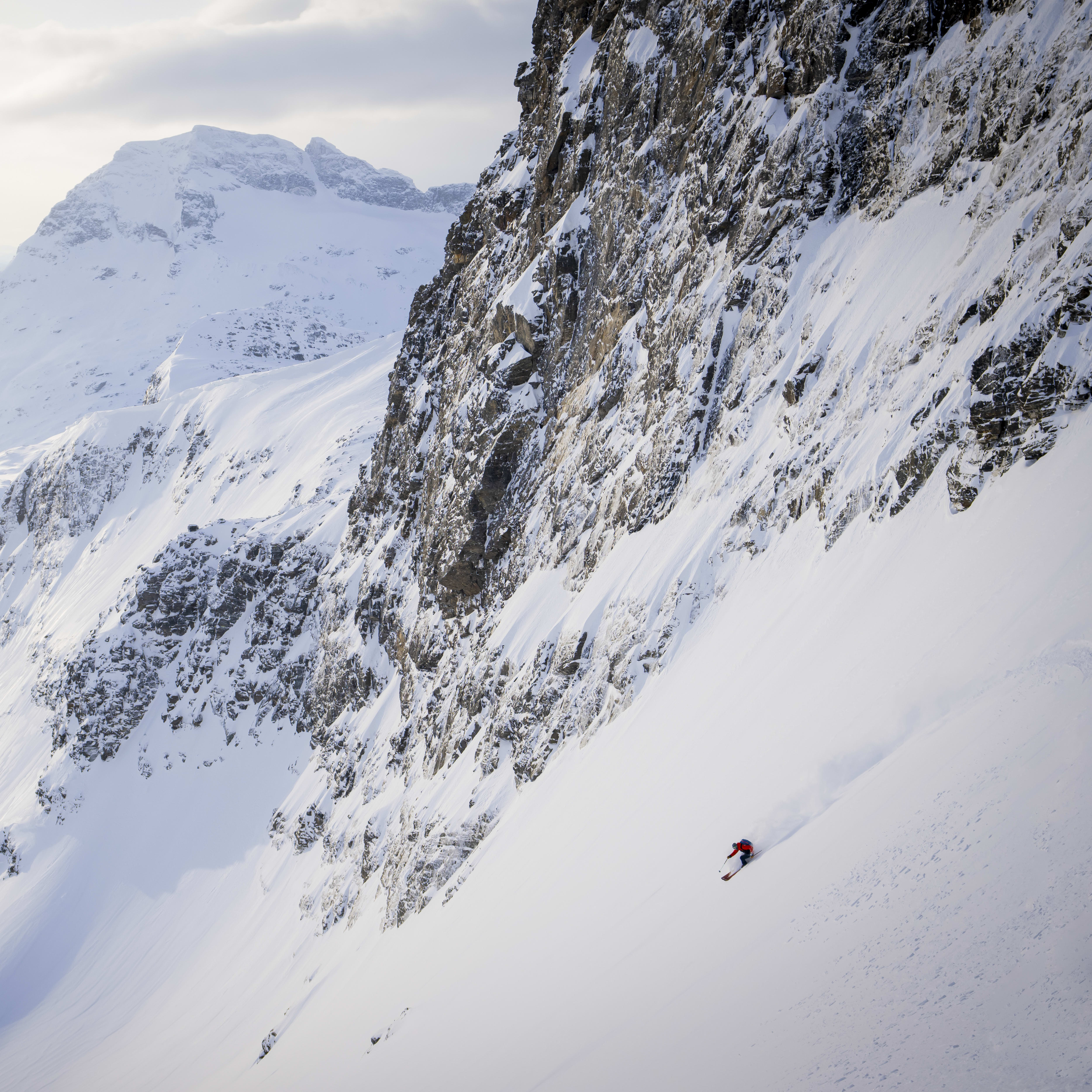 A skier in Norrøna clothing descends a steep, snow-covered mountain with rocky cliffs. Distant mountains and water are visible in the background.