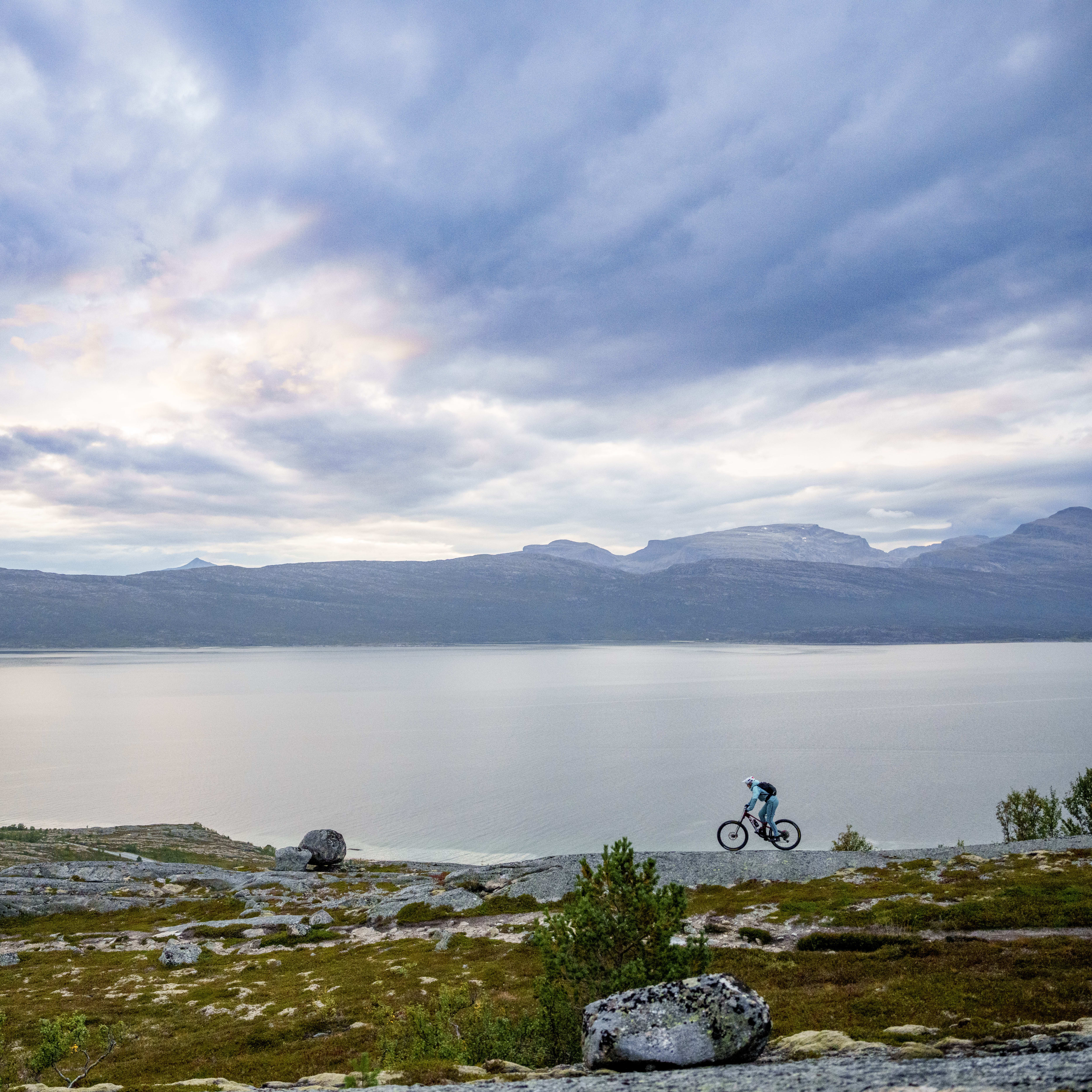 A cyclist wearing Norrøna clothing bikes along a rocky trail overlooking a calm lake and distant mountains under a cloudy sky.
