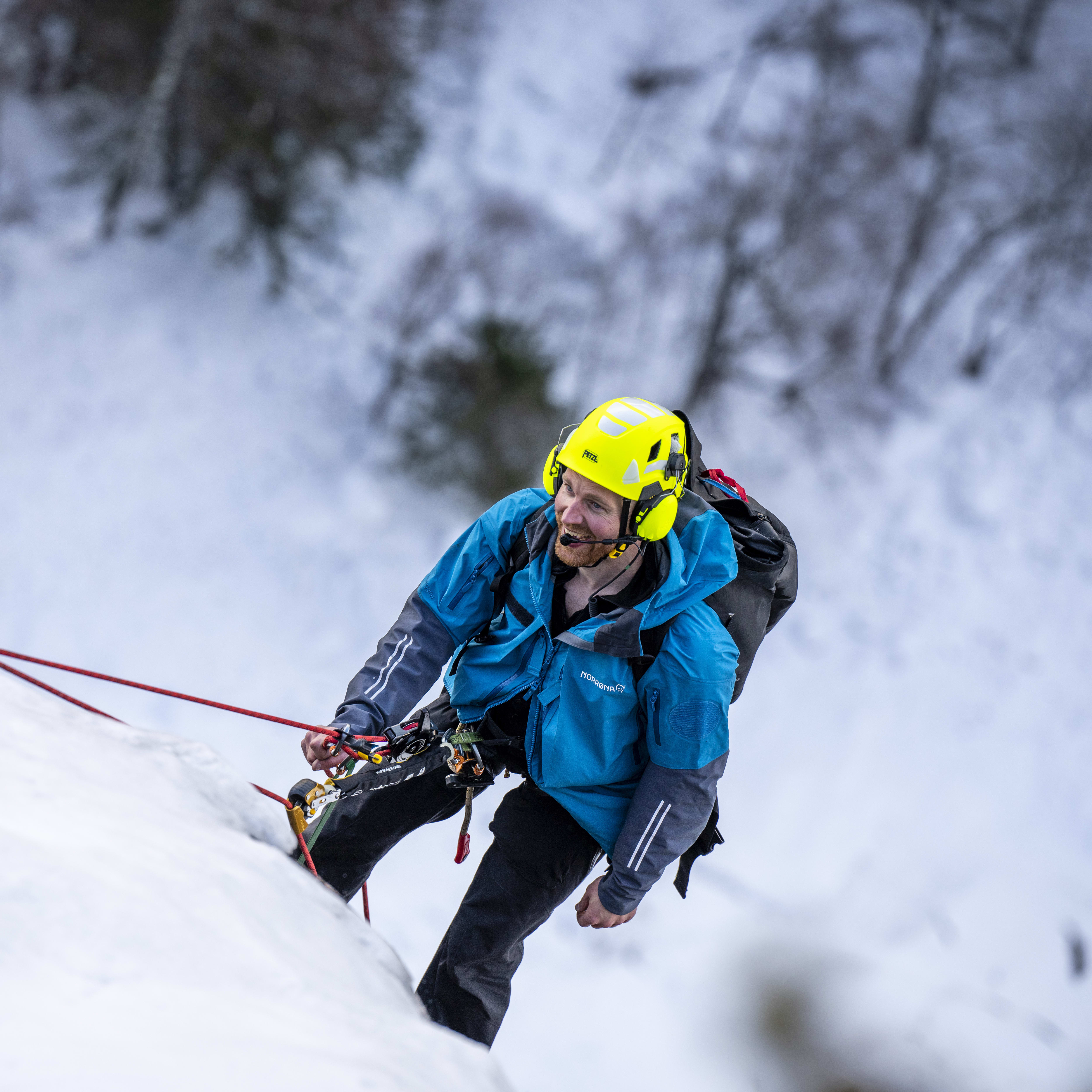 A man wearing a Norrøna jacket and a helmet climbs an icy wall. A red rope secures him to the mountain.