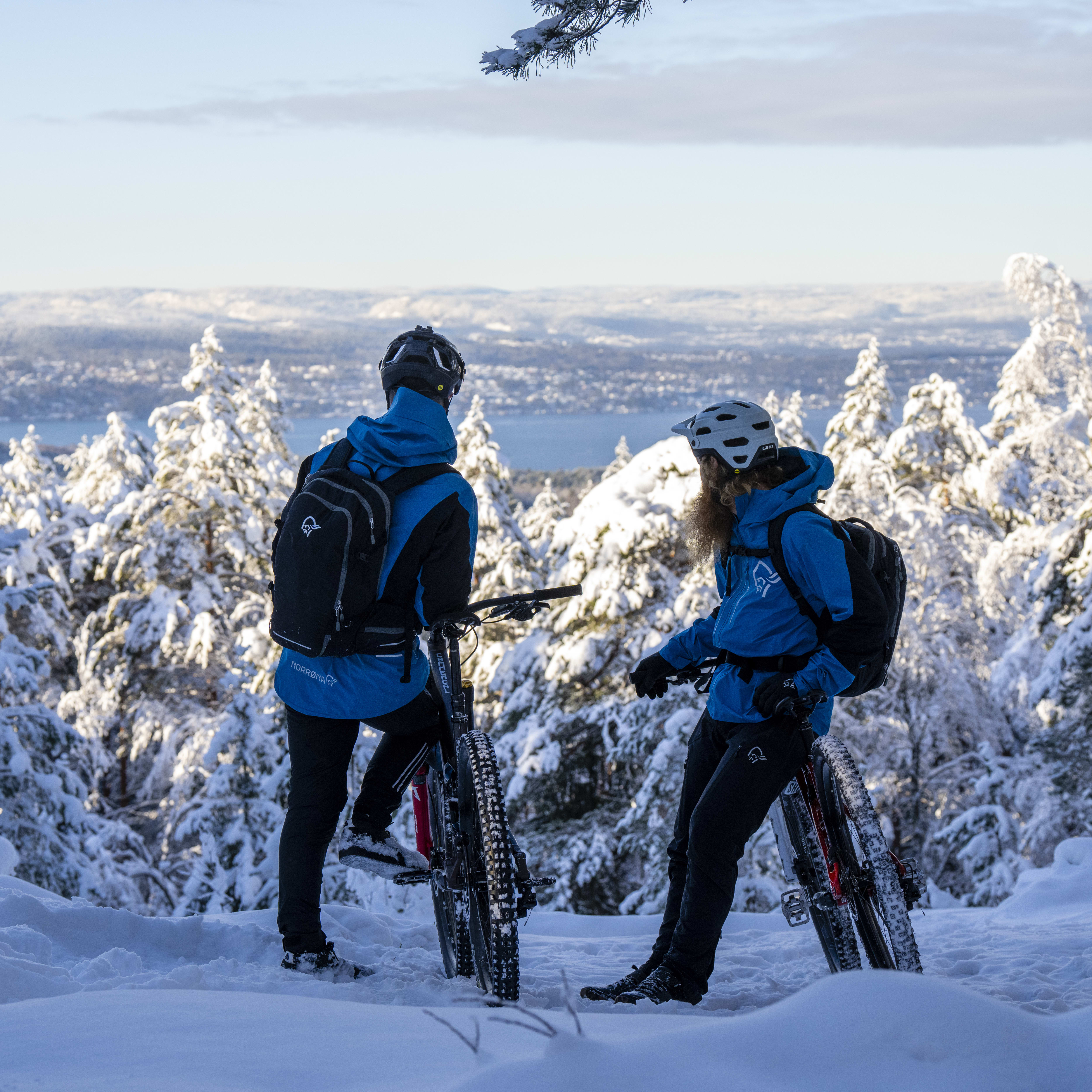 Two people in Norrøna gear stand with their bikes in a snowy forest. The background shows a mountain range and lake in the distance. 
