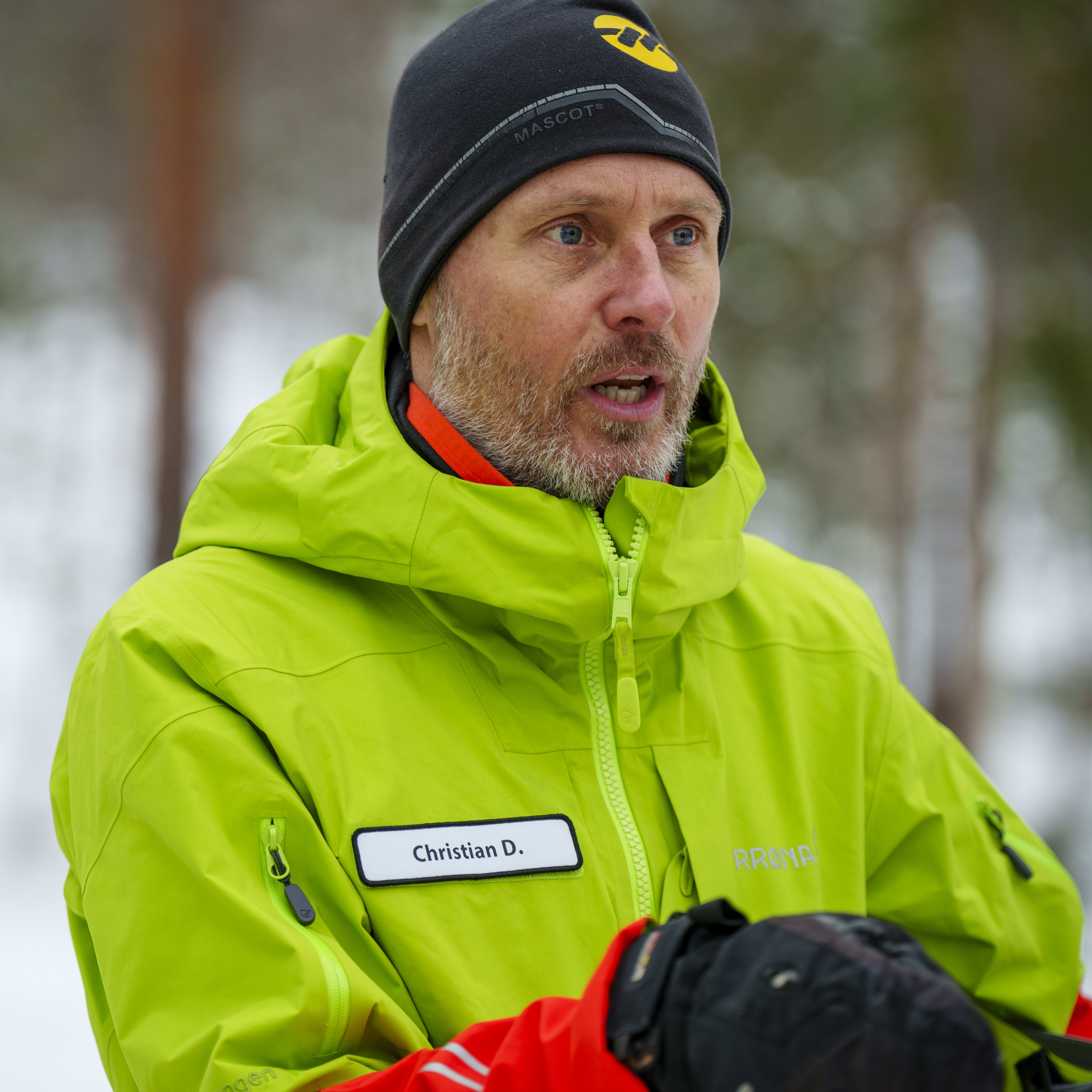 A man in a green Norrøna jacket is talking to someone off-camera. He is wearing a black hat and a name tag that reads "Christian D."  
