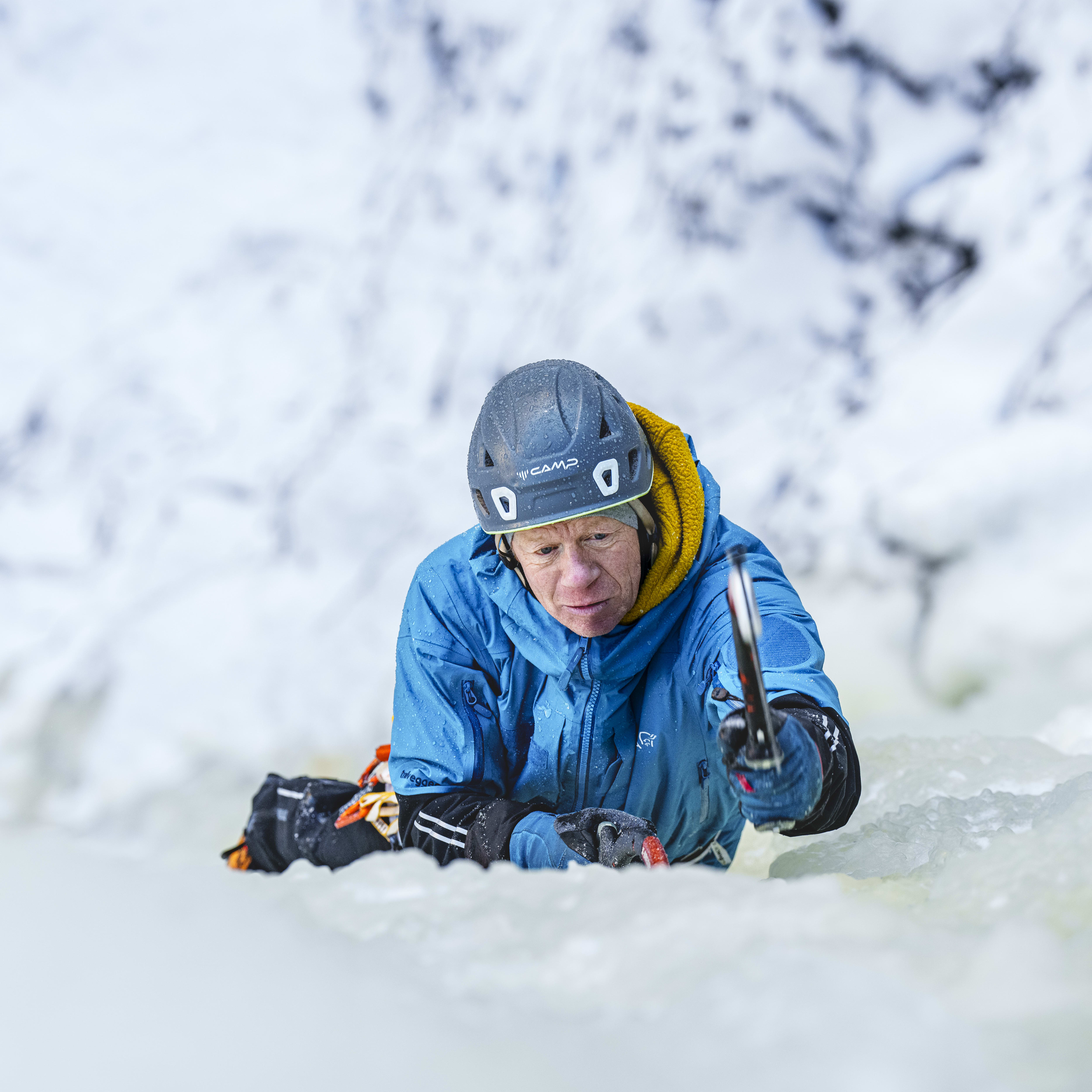 A climber wearing a blue Norrøna jacket and a helmet ascends a frozen waterfall with ice axes in hand, surrounded by snowy scenery.