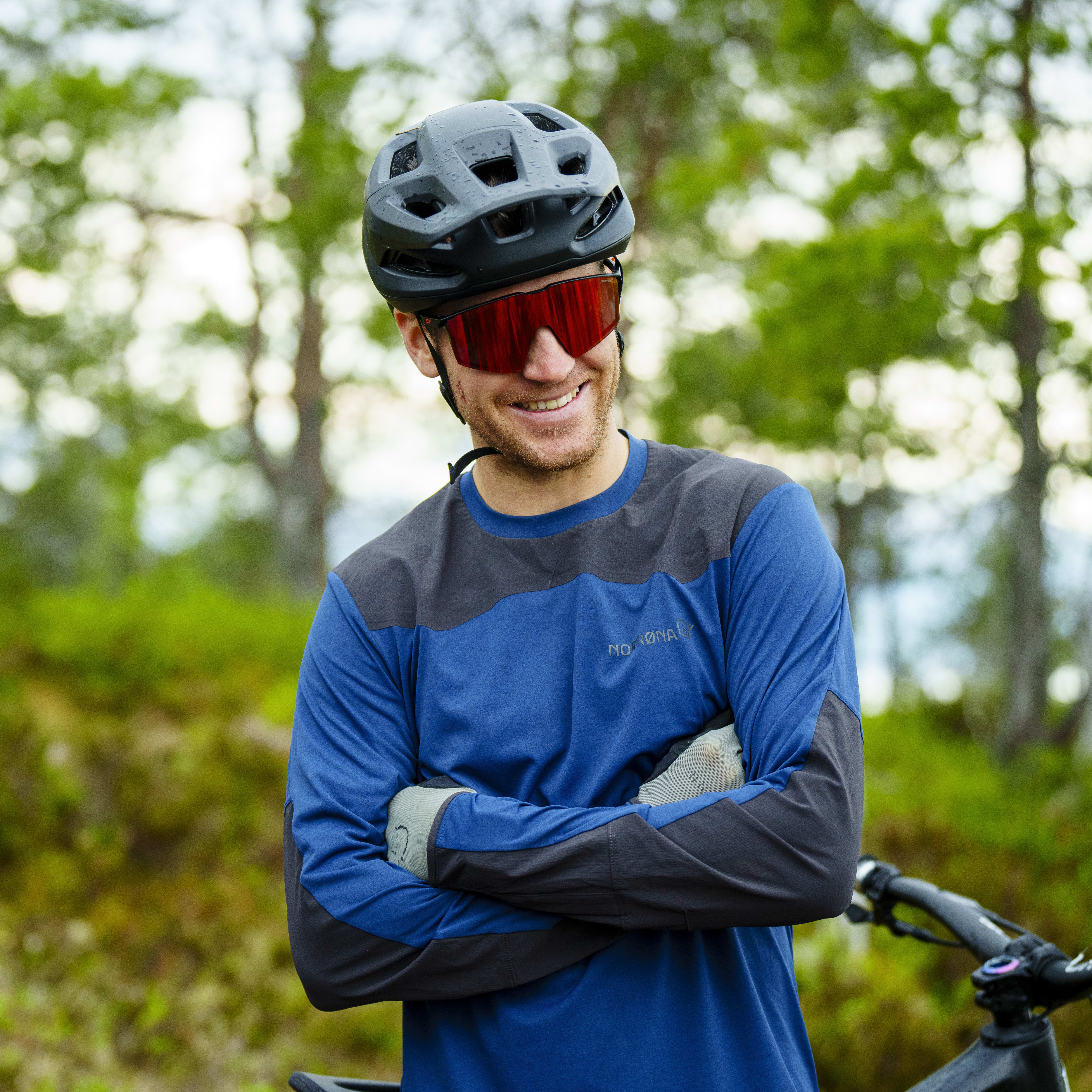 A man wearing a Norrøna long-sleeved shirt, helmet and sunglasses smiles as he stands in a forest.