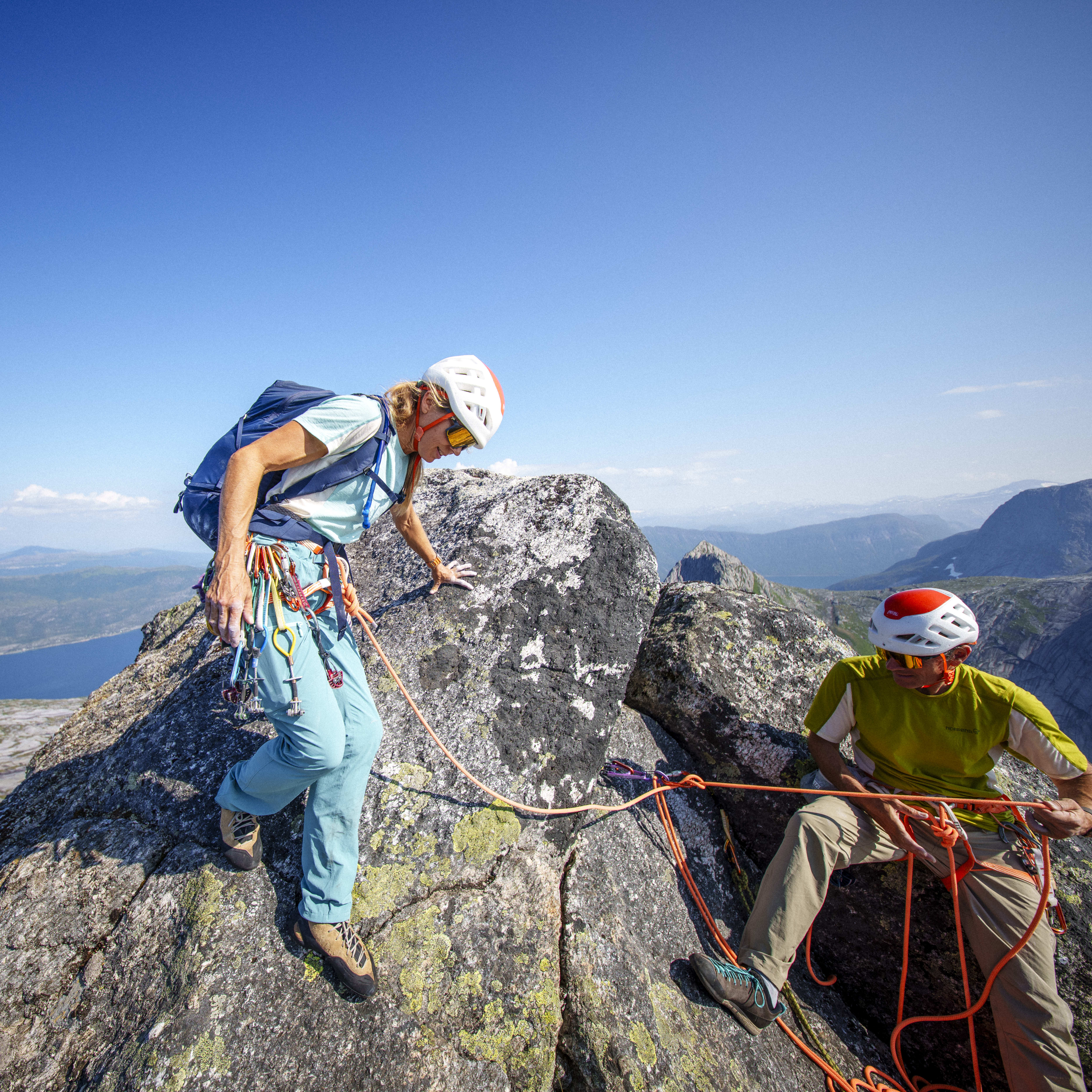 Two climbers on a rocky mountaintop against a clear blue sky and distant landscape. The woman wears Norrøna pants and a helmet. The man wears a Norrøna shirt.