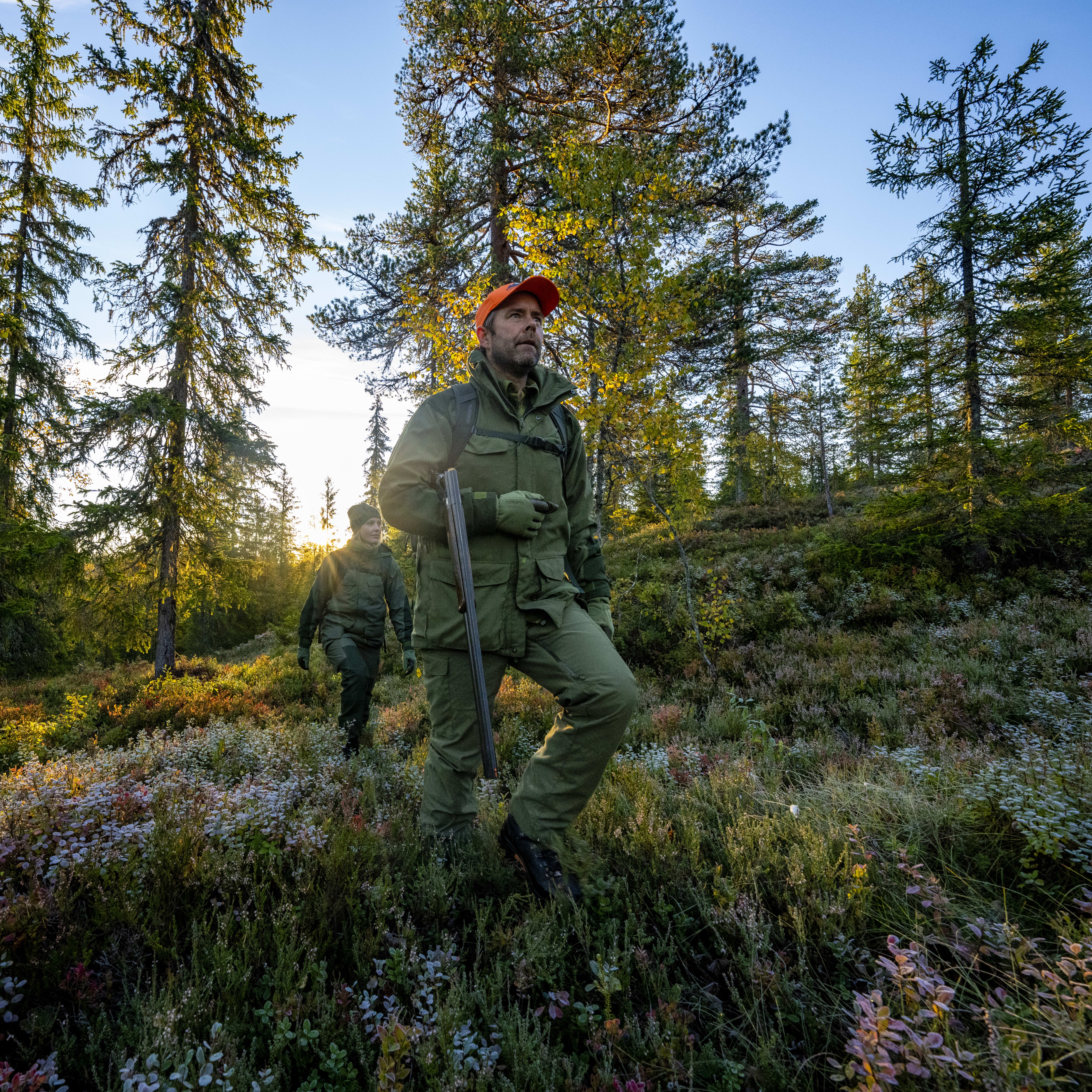 Two people wearing Norrøna clothing walk through a forest with a shotgun.  The sun shines through the trees. 

