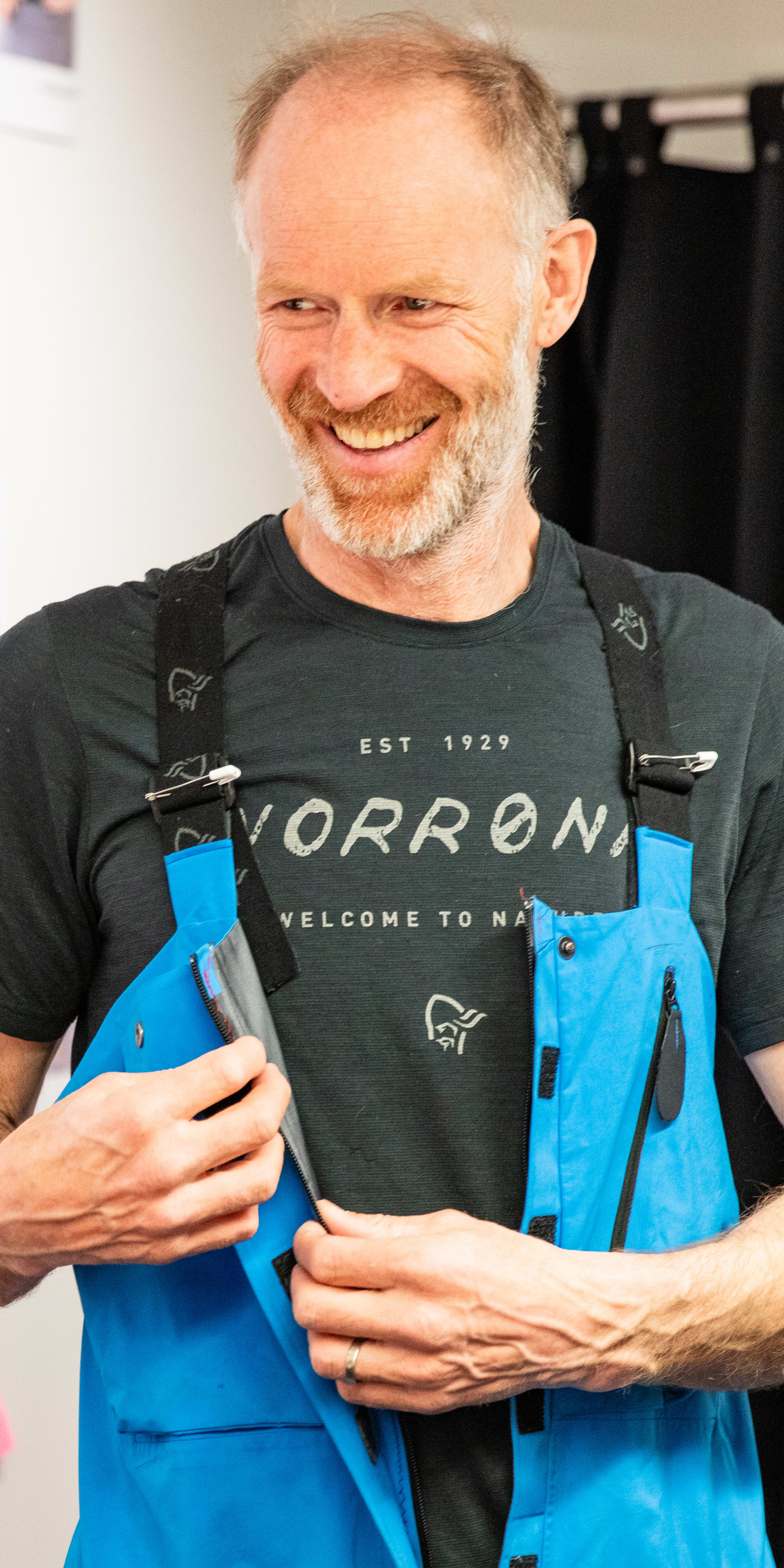 A smiling man in a black shirt adjusts a blue Norrøna jacket in front of a wall with clothing designs. 

